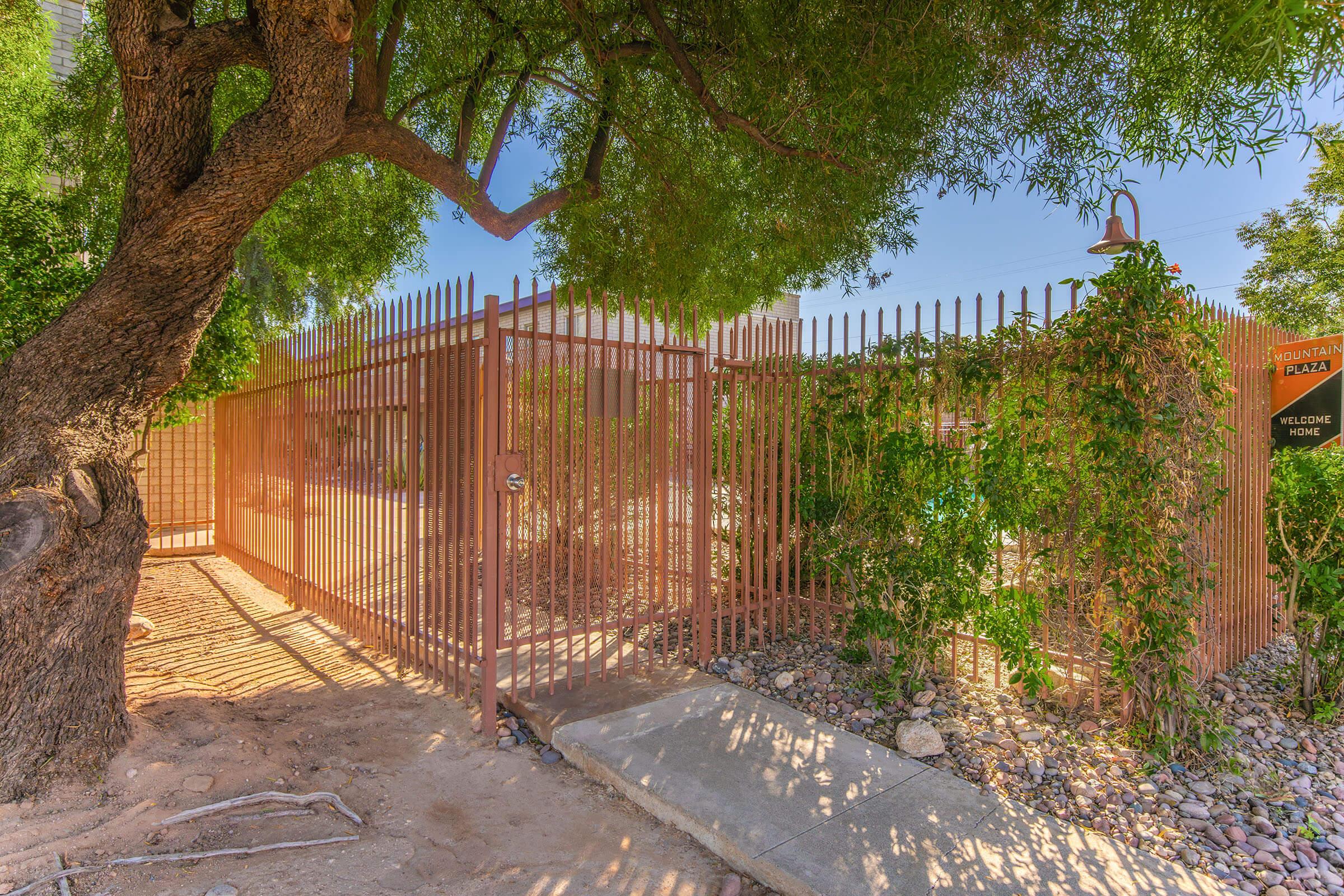 A gated entrance adorned with greenery, featuring a wrought iron fence and a prominent sign that reads "Welcome." Sunlight filters through the trees, casting shadows on the ground. The pathway leading up to the gate is lined with rocks, creating a natural and inviting atmosphere.