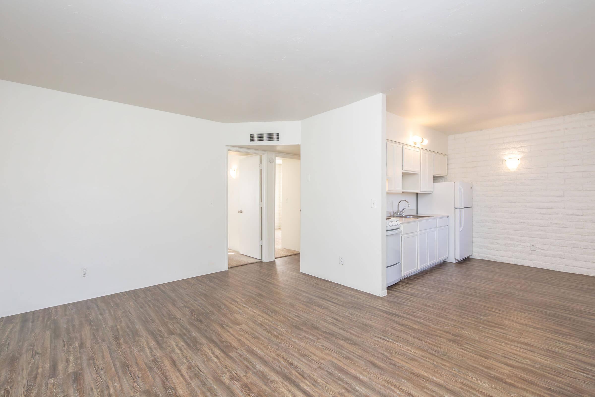 An empty, spacious living area with light-colored walls and a wooden floor. To the left, a kitchenette with white cabinets, a sink, and a white refrigerator. A doorway leads to a bathroom or bedroom, and the opposite wall features a textured panel. Soft lighting enhances the open ambiance.