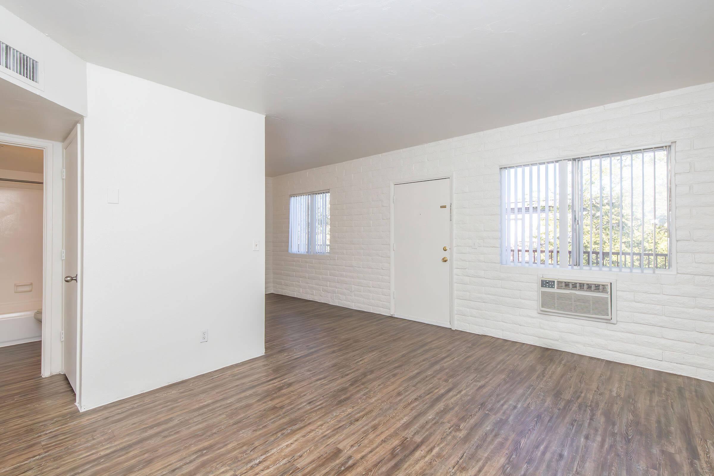 Spacious, empty living room with light-colored walls and wooden flooring. Features two vertical window units with blinds, an air conditioning unit, and a main entrance door. The design is simple and modern, creating a bright and open atmosphere.