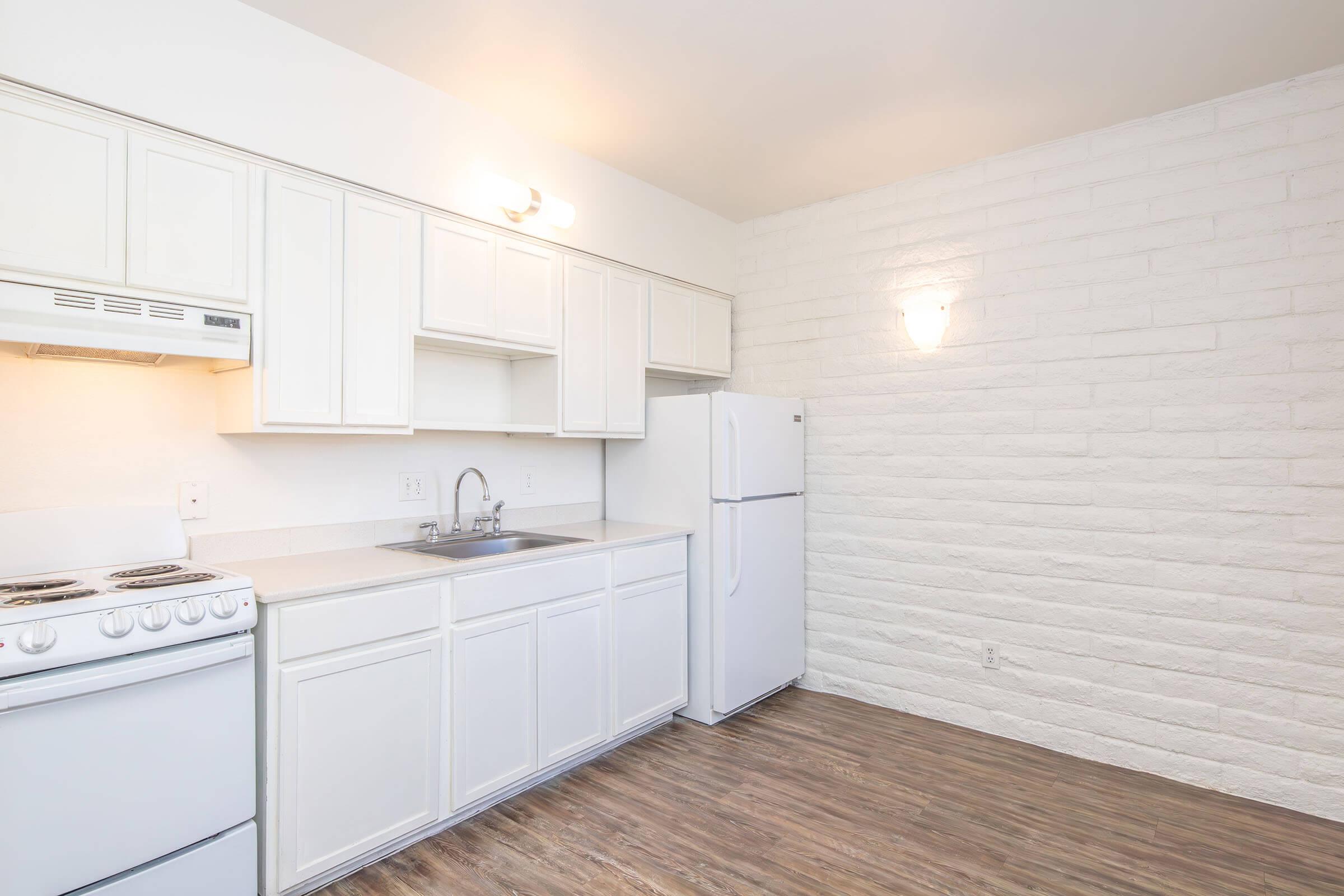 A modern kitchen featuring white cabinetry, a white stove and refrigerator, and a sink. The walls are textured with a light brick design, and the floor has a wood-like appearance. Soft lighting illuminates the space, creating a bright and clean atmosphere.