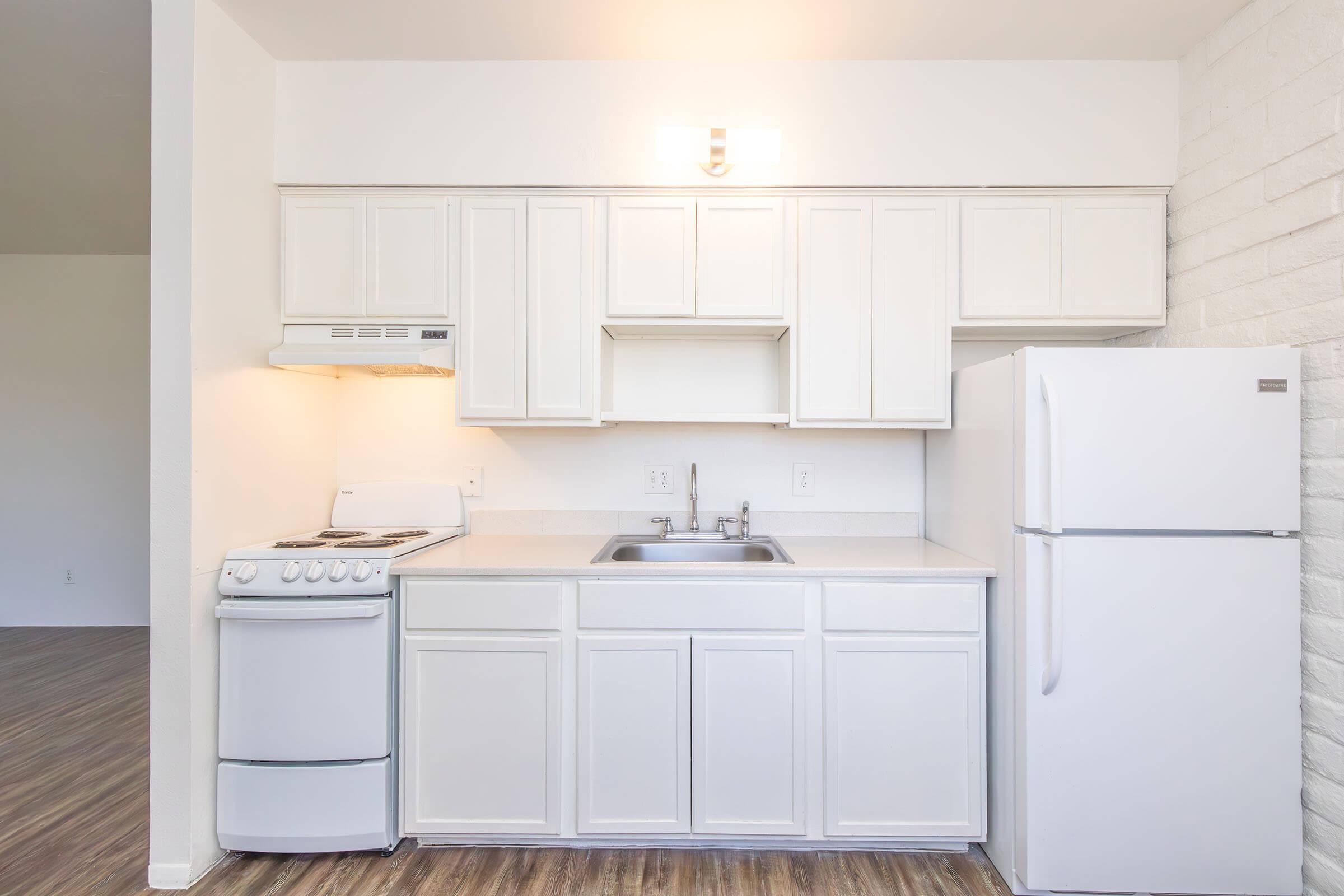 Bright, modern kitchen featuring white cabinetry, a stainless steel refrigerator, a stove with an oven, and a sink. The kitchen has a clean, minimalist design with light-colored walls and wooden flooring, creating a spacious and inviting atmosphere. A wall-mounted light fixture provides additional lighting.