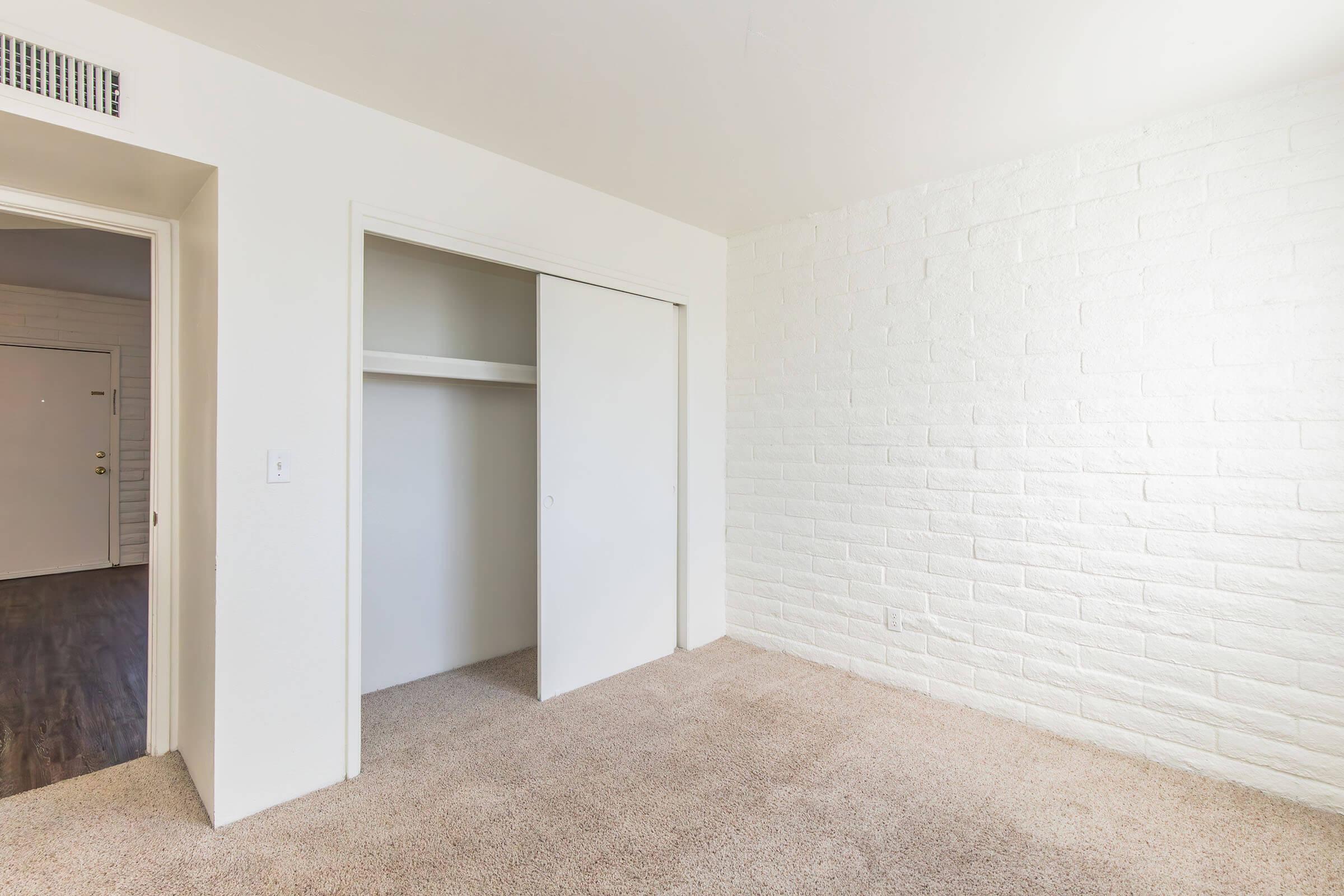 A spacious room with light-colored walls and a textured white brick feature wall. The floor is covered in beige carpet, and a closet with sliding doors is visible on the left. The entrance door can be seen in the background, suggesting a welcoming and bright interior space.