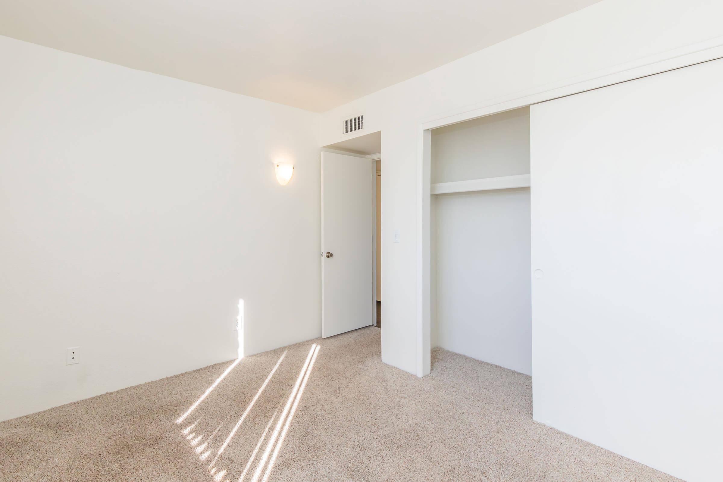 A simple, empty bedroom featuring neutral-colored walls and carpet. The room has a single light fixture on the wall, a closed door leading to another space, and a white closet with sliding doors. Light streams in through a window, creating subtle shadows on the carpeted floor.