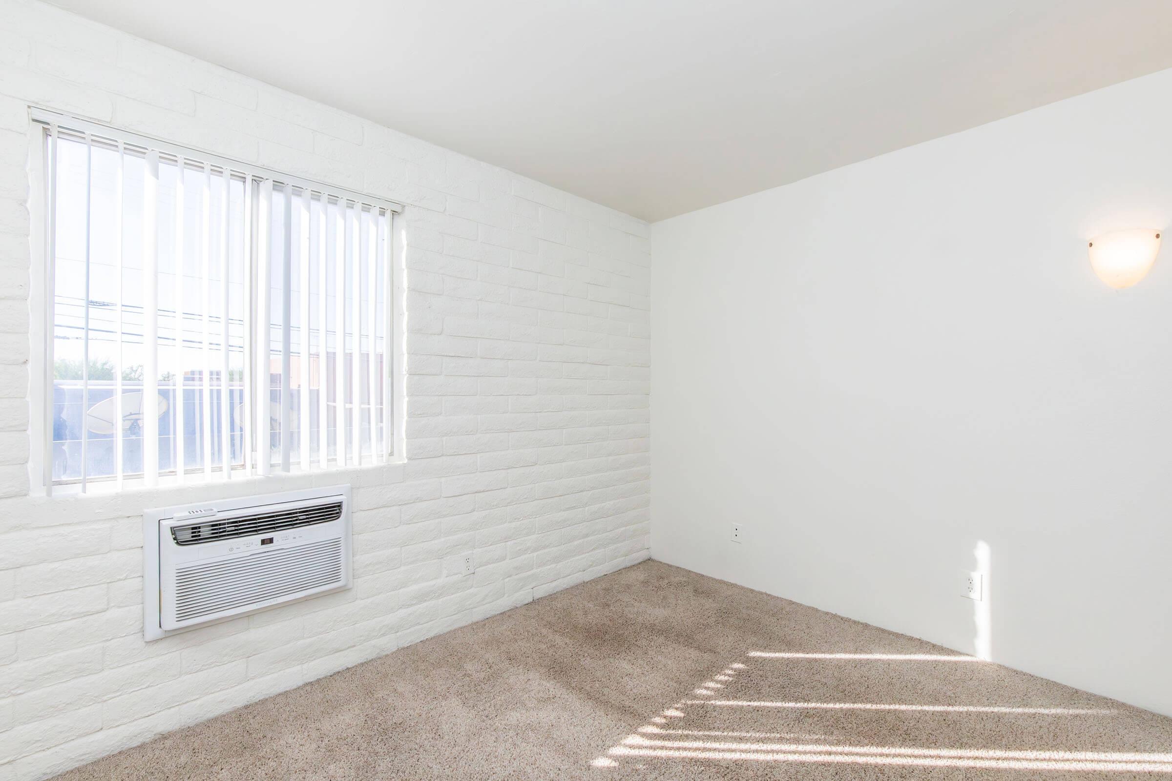 Empty room with white walls and a light beige carpet. There is a window with vertical blinds allowing natural light in, and an air conditioning unit beneath the window. The overall space is bright and minimalistic, featuring a single wall-mounted light fixture.