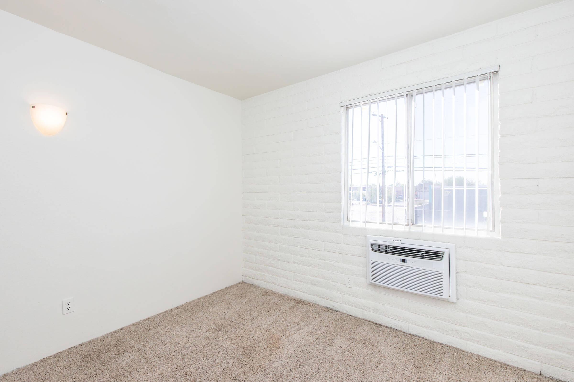 A bright, empty room with white brick walls, featuring a window with vertical blinds and an air conditioning unit below it. The floor is covered in light-colored carpet, and there is a wall-mounted light fixture. Overall, the space has a clean and minimalist appearance.