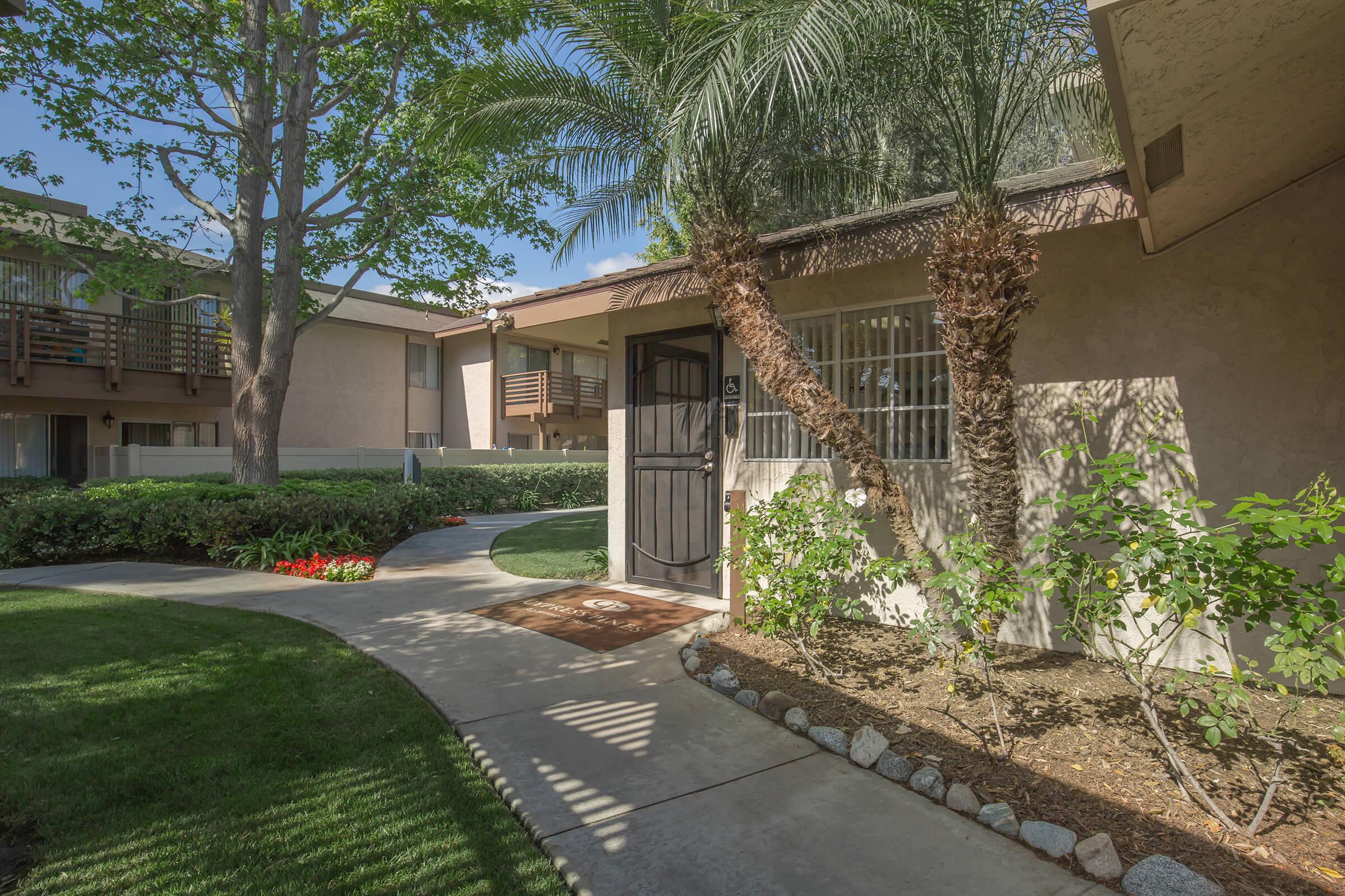 A scenic view of a landscaped path leading to an entrance of an apartment with palm trees and greenery surrounding the area. Flower beds and well-maintained lawns enhance the inviting atmosphere of the residential complex.
