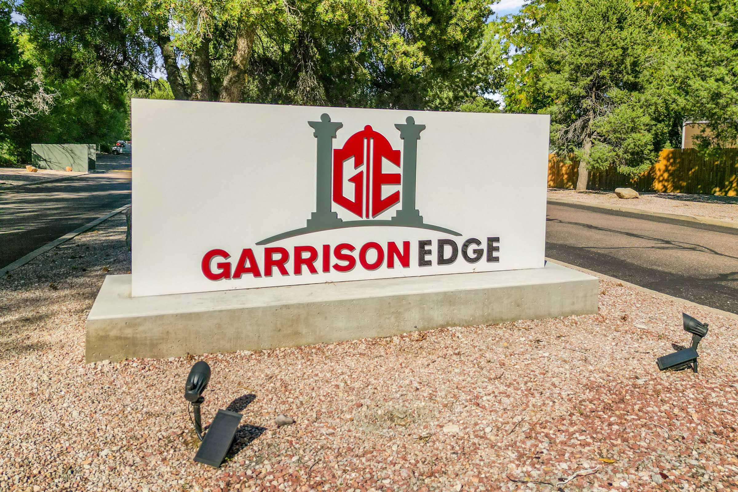 Sign for "Garrison Edge" featuring the letters "GE" in red and a stylized design of pillars. The sign is set in a landscaped area with trees and gravel ground, indicating a welcoming entrance to the facility or community.