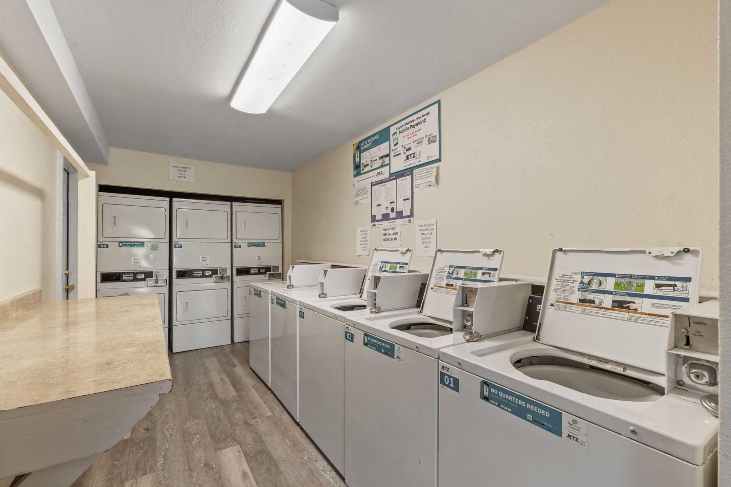 A clean and modern laundry room featuring several white washing machines and dryers against a beige wall. There is a countertop for folding clothes and informational posters on the wall. The space is well-lit with fluorescent ceiling lights, creating a bright and inviting atmosphere.