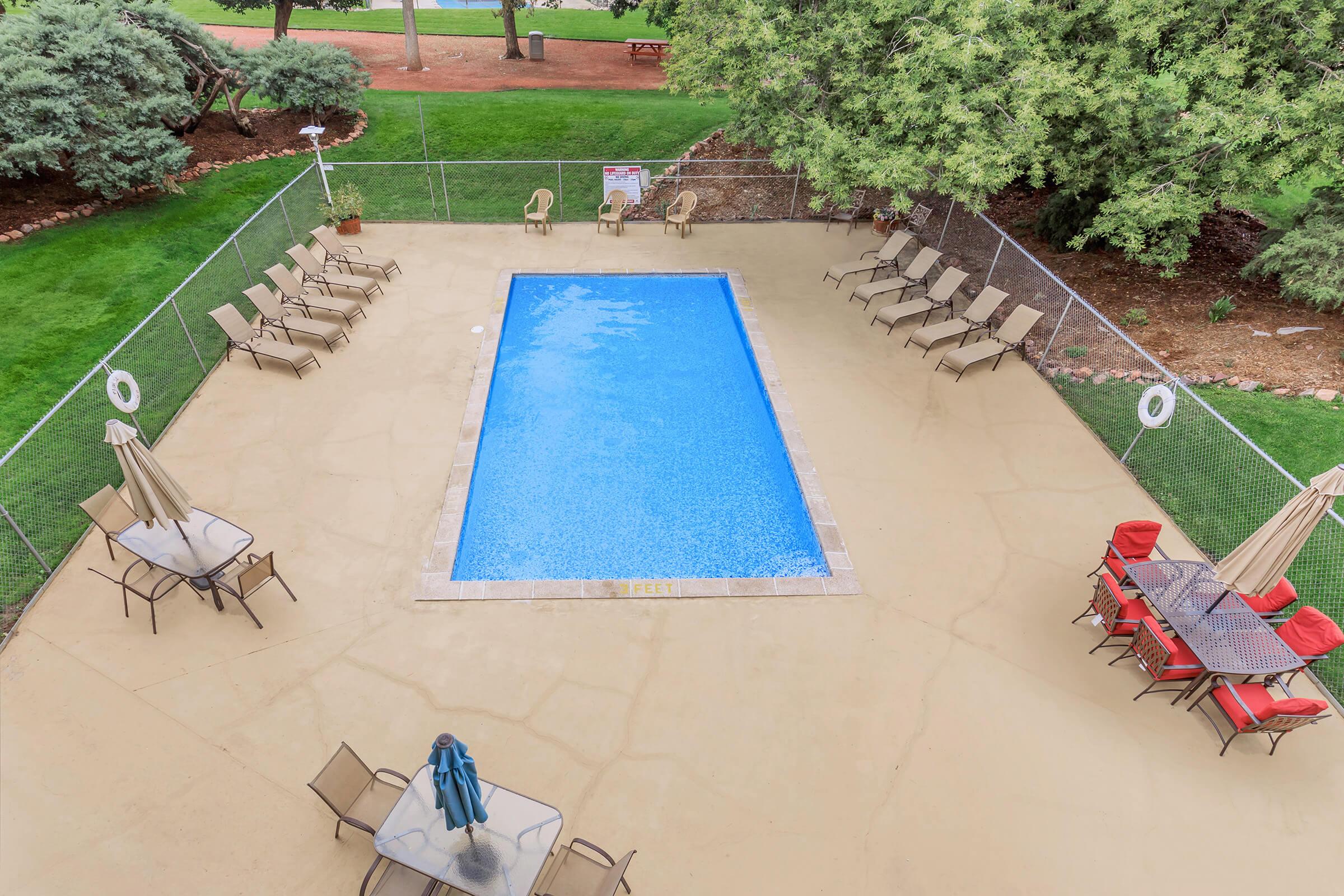 Aerial view of a fenced swimming pool area surrounded by green trees. The pool is clear blue and rectangular, with several lounge chairs arranged around it. There's a small table set with red chairs nearby and umbrellas for shade. The surrounding area is landscaped with grass and shrubs.