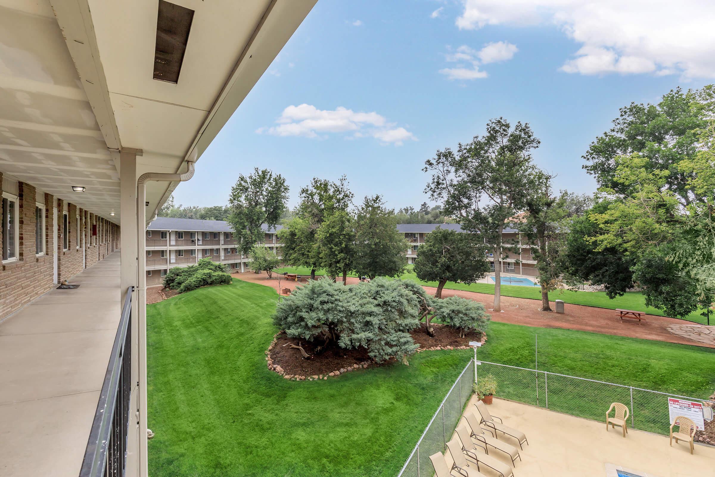 View from a balcony overlooking a manicured green lawn with trees and shrubs, surrounded by two-story buildings. A swimming pool is visible in the background, and there are chairs and tables in the outdoor area. The sky is partly cloudy, adding to the serene atmosphere.