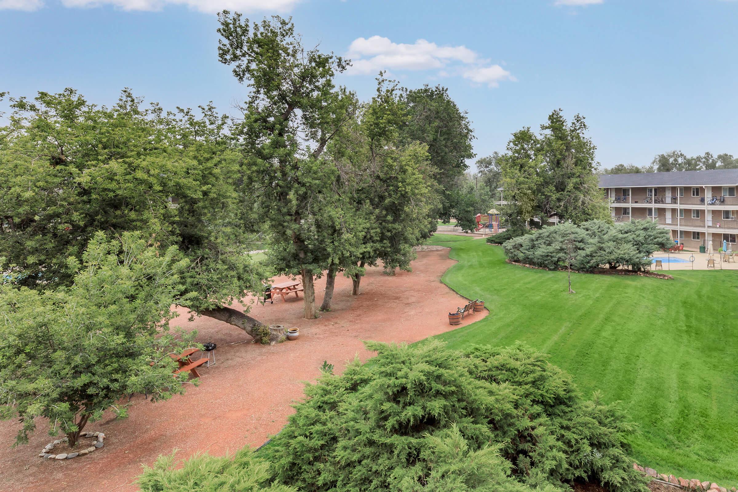 A tranquil outdoor area featuring green grass and trees, with a pathway meandering through. There are picnic tables arranged under the trees, and a low brick wall separates the space from a building in the background. The sky is clear, contributing to a peaceful, inviting atmosphere.