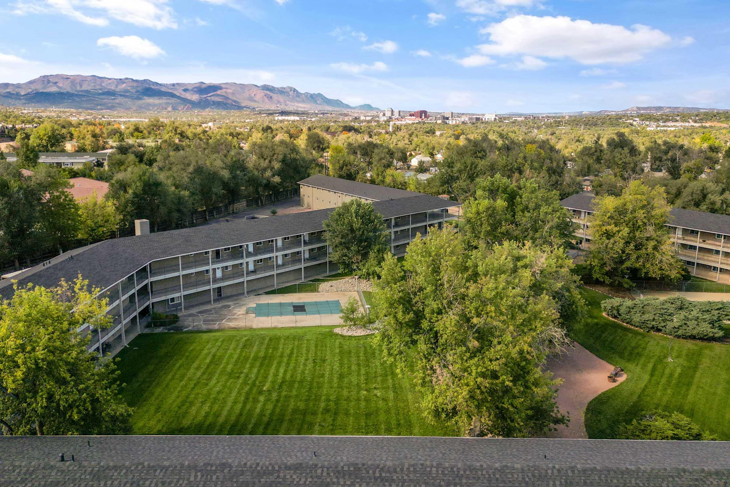 Aerial view of a multi-story hotel with a courtyard and green lawn surrounded by trees. Mountains are visible in the background, along with a distant view of a city skyline under a blue sky with scattered clouds.