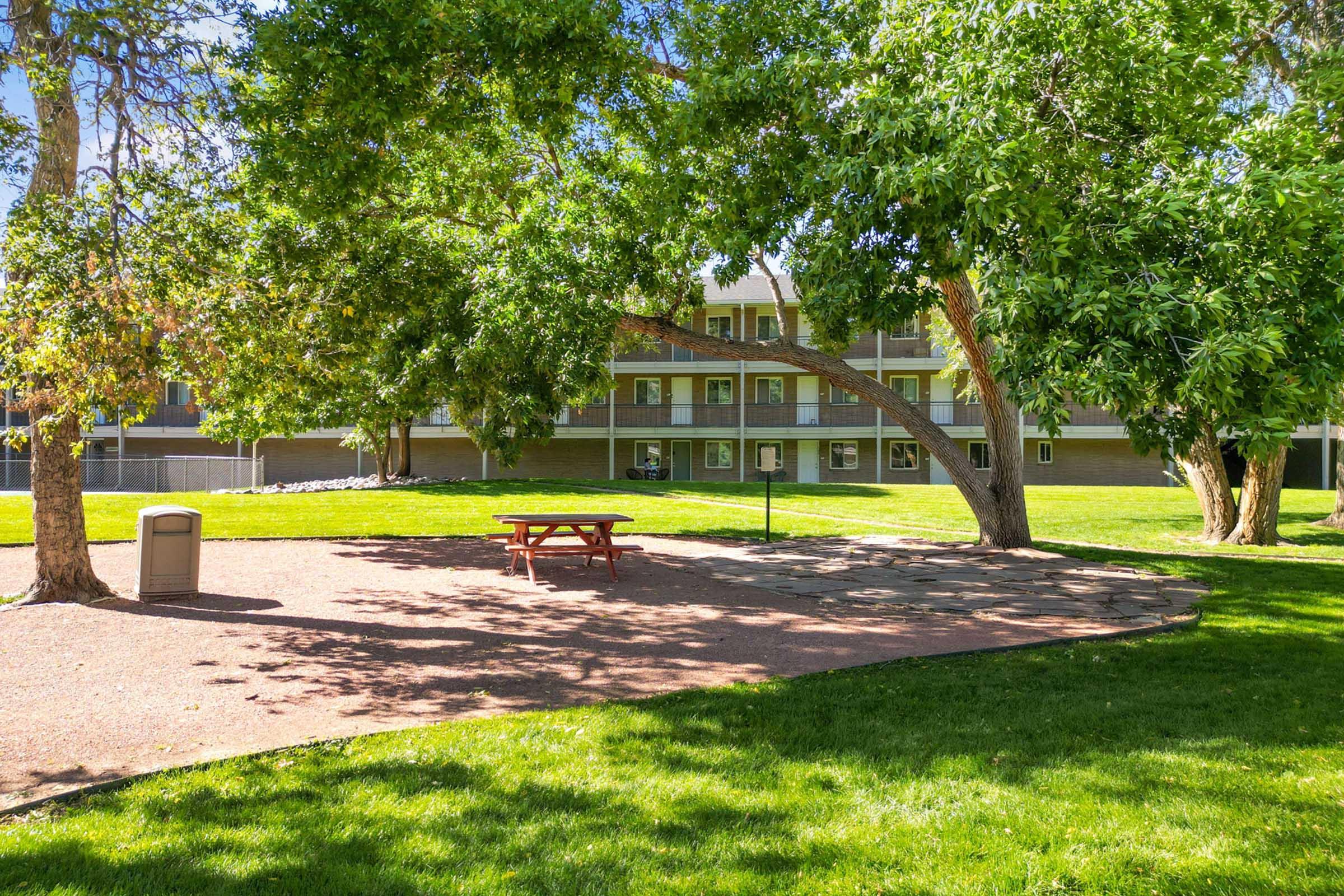A grassy area with a picnic table in the foreground, surrounded by trees. In the background, there is a multi-story building with balconies. The scene is sunny, with clear skies and a well-maintained lawn, creating a peaceful outdoor space.
