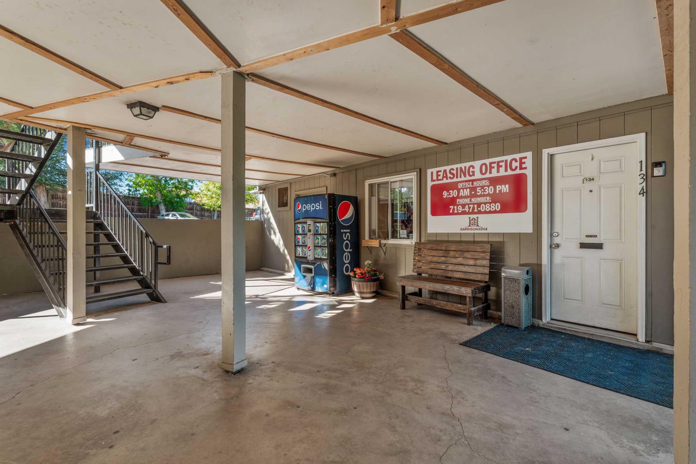 A covered leasing office area with a Pepsi vending machine, a bench, and stairs leading to an upper level. The office door is labeled with hours of operation. The space is well-lit and features concrete flooring.