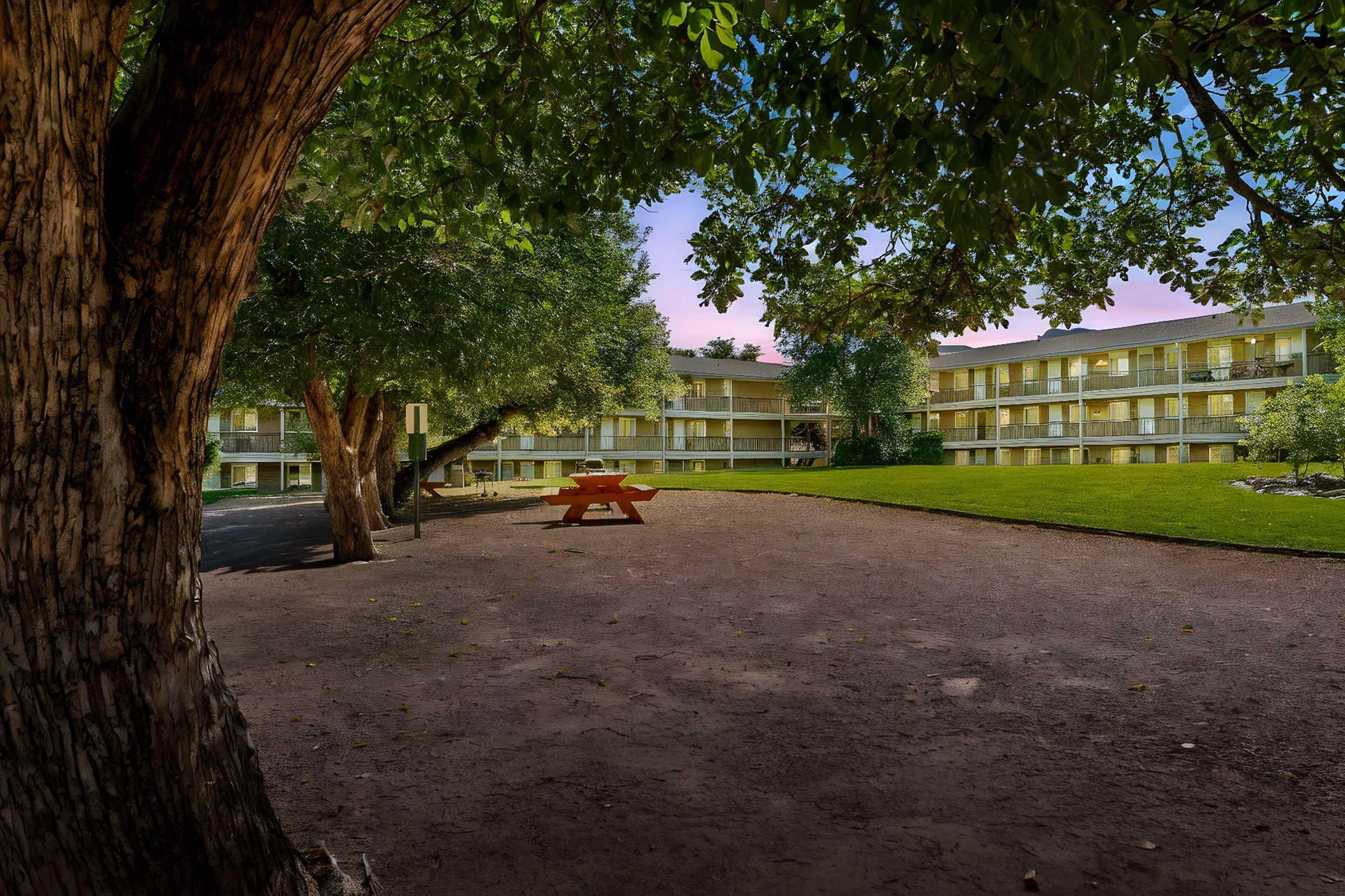 A peaceful courtyard scene featuring a large tree on the left, with a grassy area and a picnic table. Surrounding the courtyard is a two-story building with multiple balconies. The sky displays soft pastel hues, indicating sunrise or sunset, creating a tranquil atmosphere.