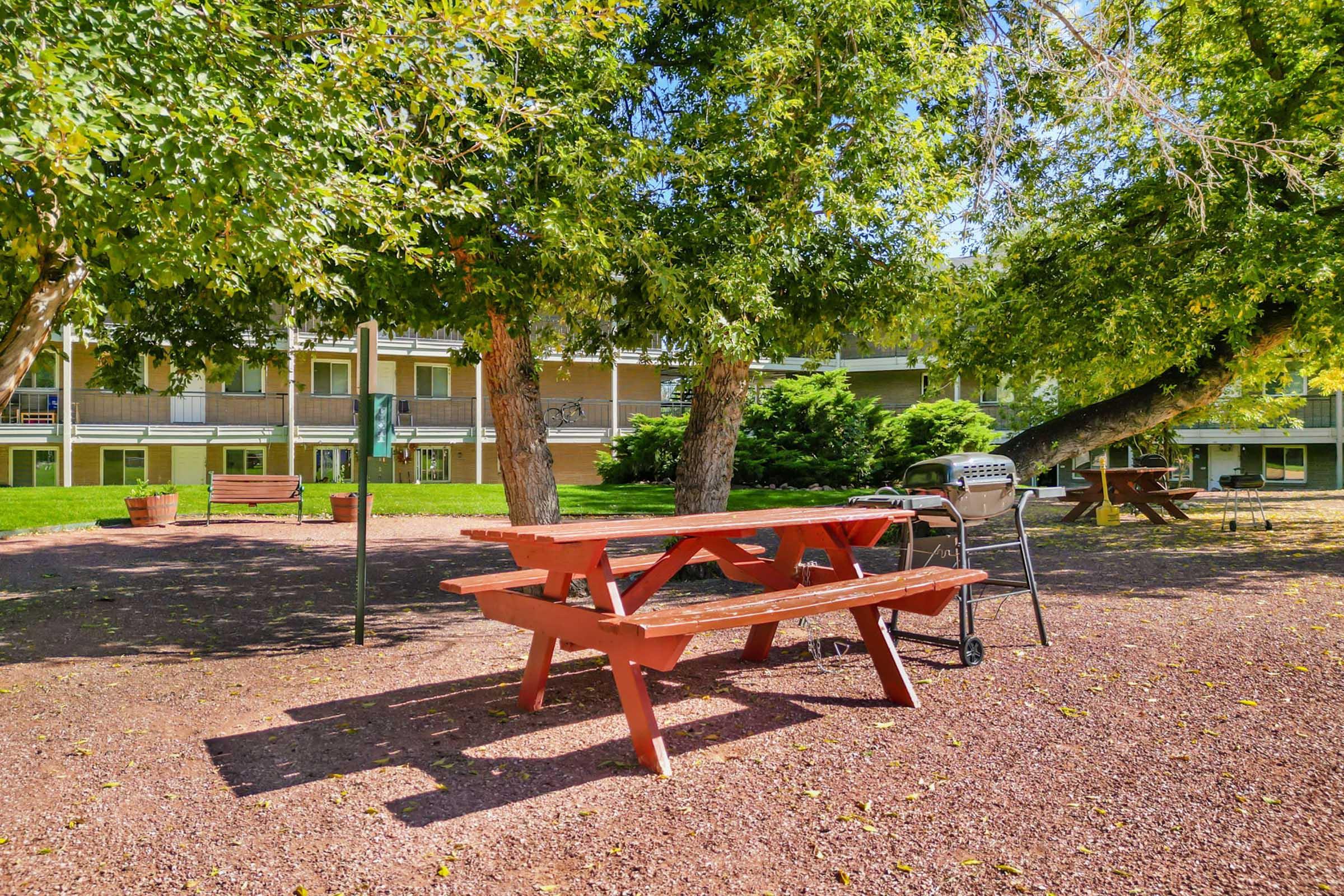 A red picnic table is set in a shaded area surrounded by green trees. In the background, there are multiple buildings with balconies. The ground is covered with small stones, and there’s a grill nearby. The atmosphere is serene, suggesting a peaceful outdoor space for relaxation.