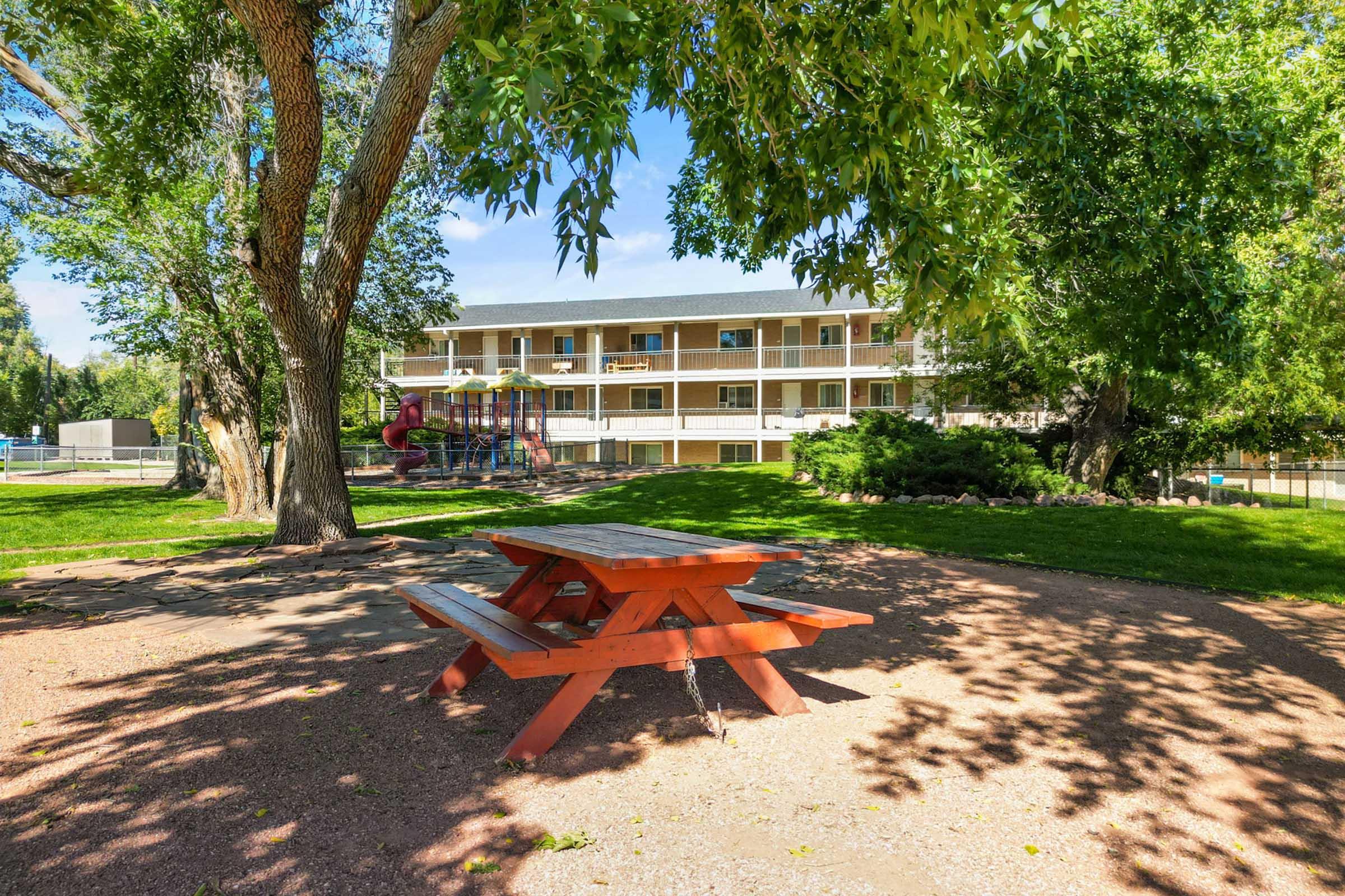A picnic table sits under a tree in a grassy area, with a playground visible nearby. In the background, a two-story building with balconies can be seen against a clear blue sky. The scene conveys a peaceful outdoor setting suitable for relaxation and recreational activities.