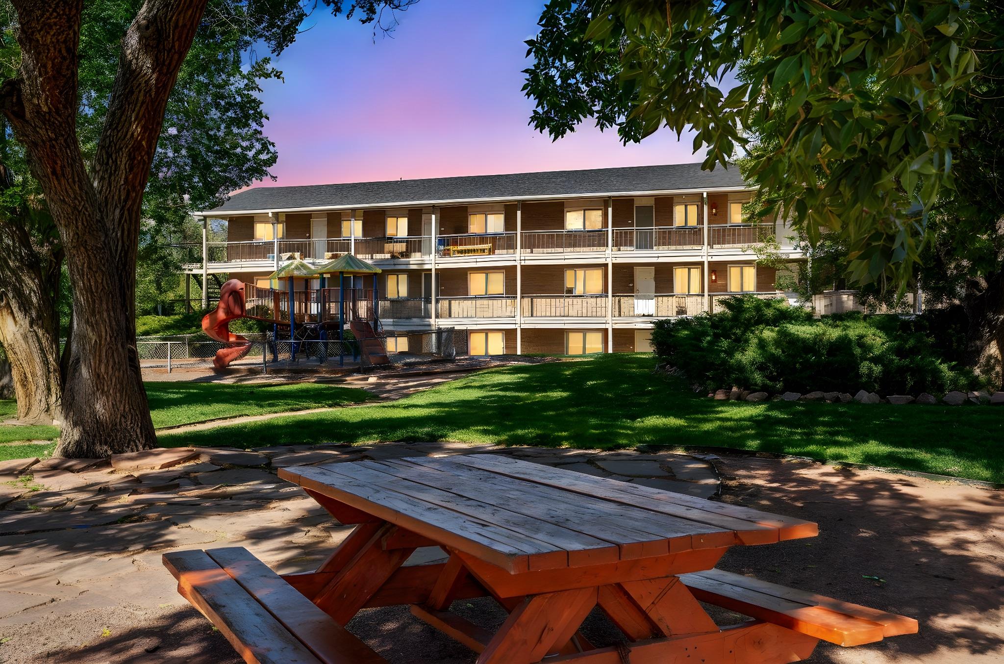 A wooden picnic table in a grassy area in front of a two-story building with multiple balconies. The building features large windows reflecting the surrounding trees and a colorful playground with a slide nearby. The sky above has a gradient of colors, suggesting early morning or late afternoon light.