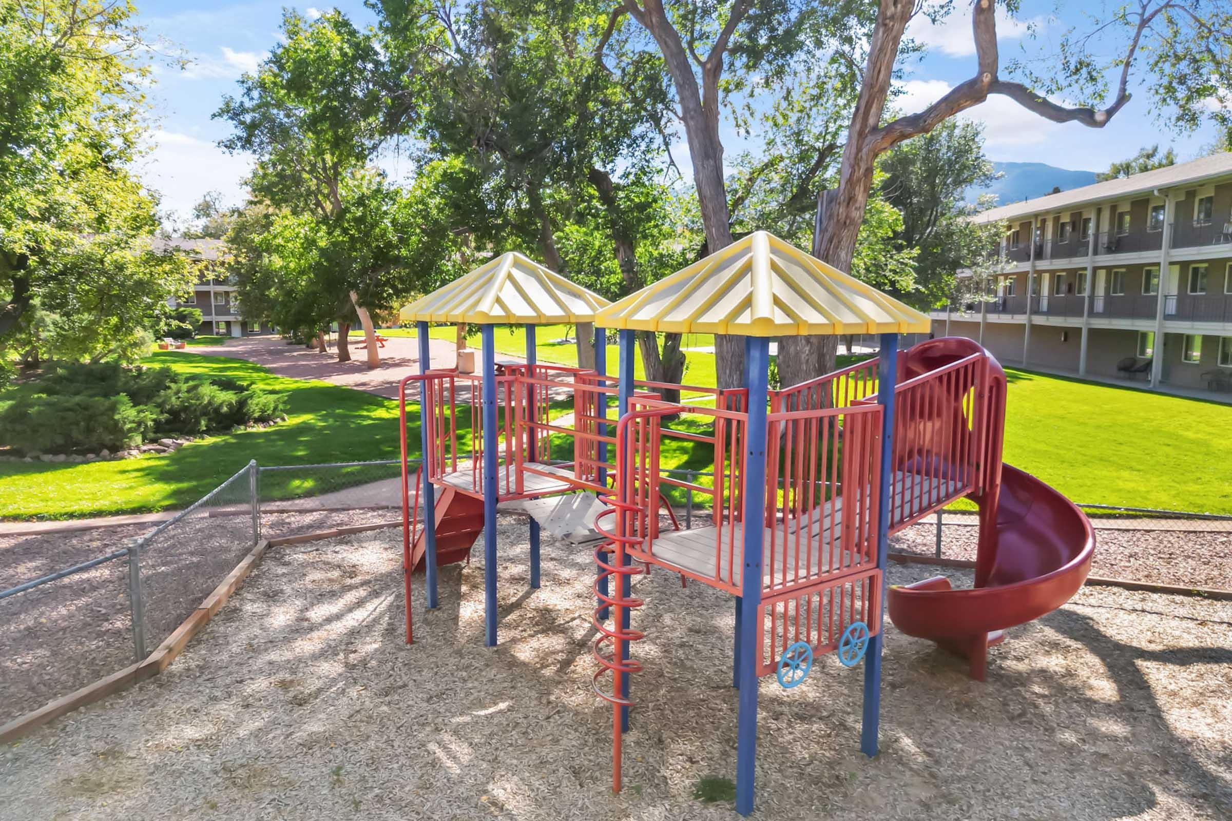 Playground with colorful play structures, featuring two towers with yellow and red roofs, a spiral slide, and a larger red slide. Surrounding grassy area with trees and an apartment building in the background. Bright, sunny day with clear blue skies.
