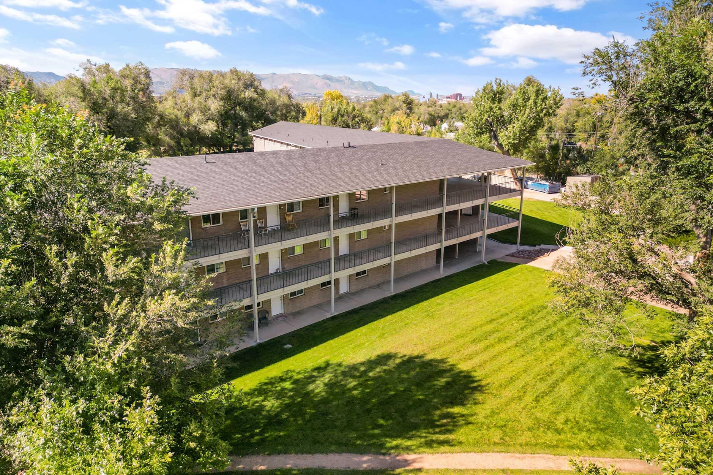 Aerial view of a modern apartment building surrounded by trees and green grass. The building features multiple balconies and a landscaped area in front, with a swimming pool visible in the background. The scene is bright and sunny, showcasing a pleasant residential environment.
