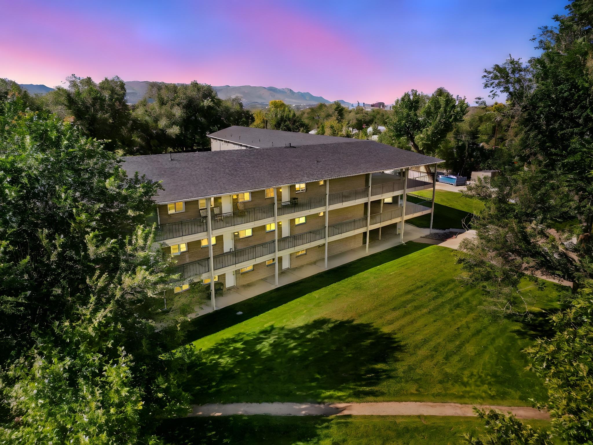 A three-story building surrounded by lush greenery and trees, with a pathway leading through the grass. The sky is painted in shades of pink and blue, creating a serene evening atmosphere. The building features several balconies with illuminated windows, indicating activity inside.
