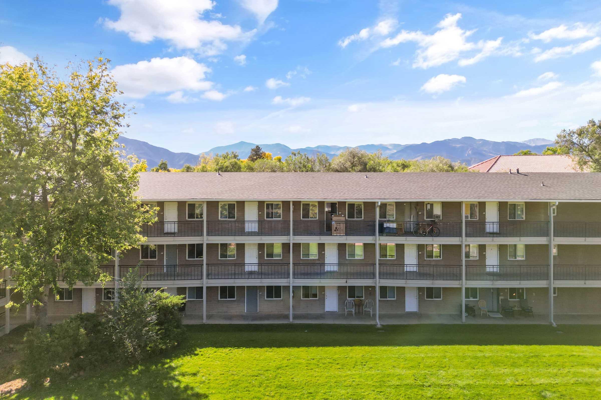 A view of a multi-story apartment building with balconies, set against a backdrop of mountains and a clear blue sky. The foreground features a green lawn with trees, and some furniture can be seen on the balconies, indicating a residential environment.