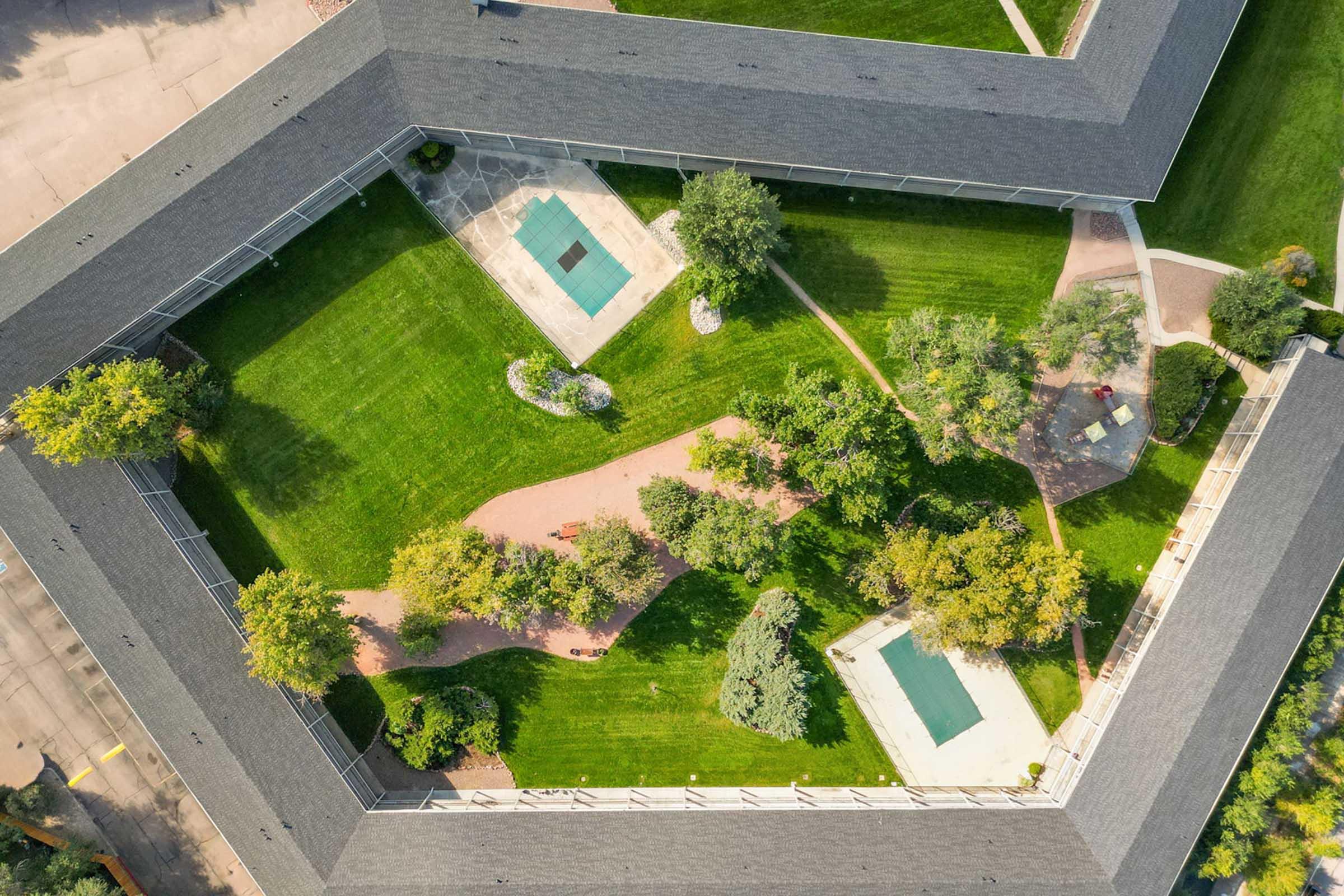 Aerial view of a landscaped courtyard featuring green lawns, walking paths, and small trees. Two rectangular pools are visible, along with garden features and seating areas, all enclosed by buildings. The layout creates a serene, inviting environment.