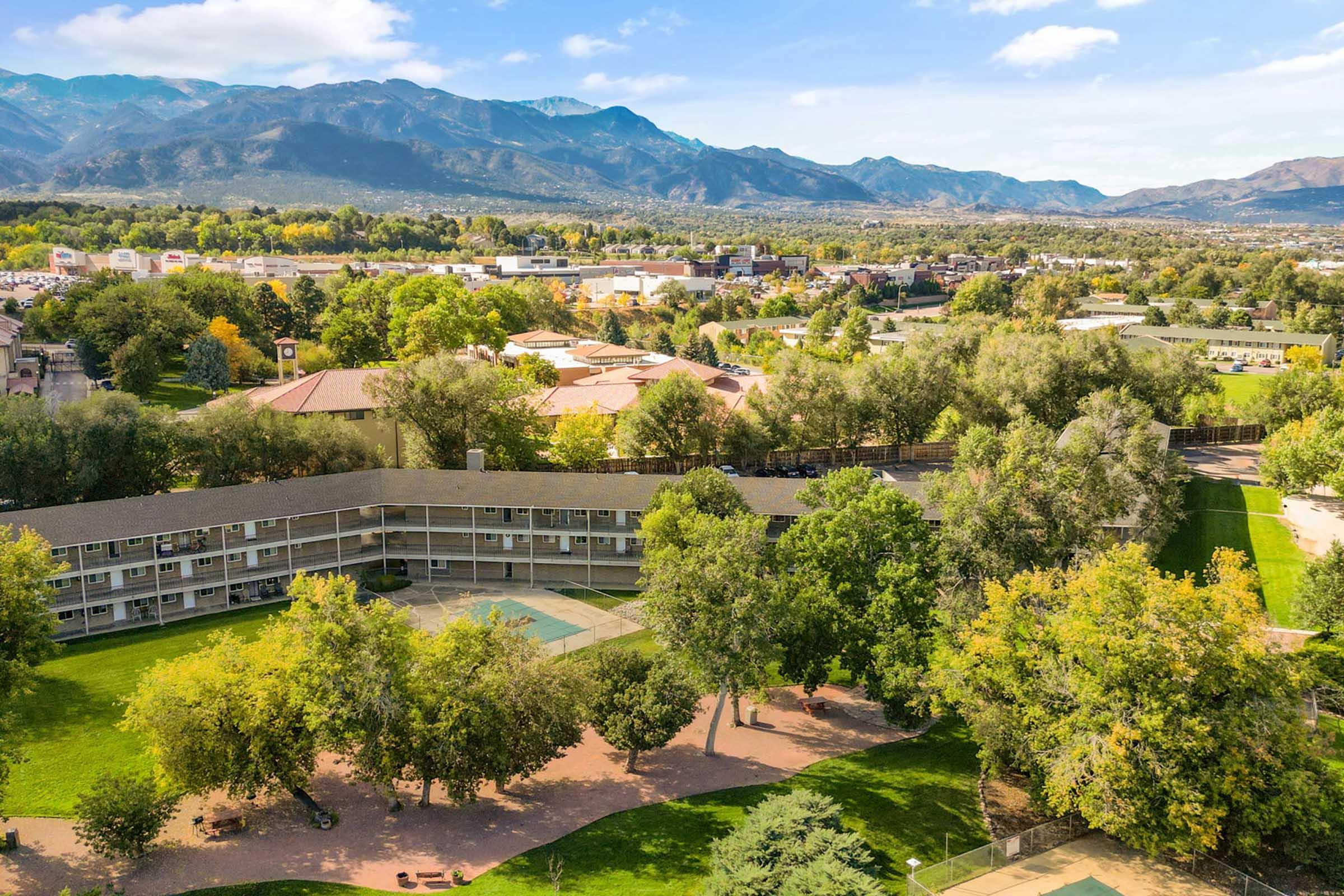 An aerial view of a hotel with a courtyard surrounded by trees and green grass, set against a backdrop of mountains. The landscape features a mix of residential and commercial buildings in the distance, with clear blue skies and scattered clouds.