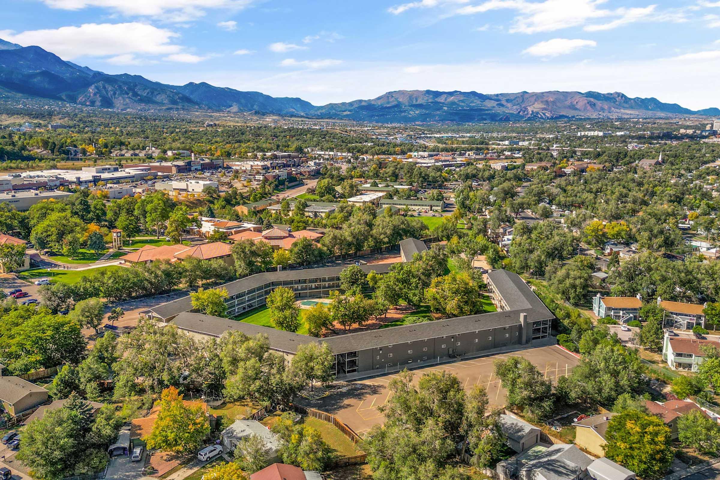 Aerial view of a vibrant landscape featuring clusters of greenery, residential buildings, and a commercial area, against a backdrop of mountains and a blue sky with scattered clouds. The scene captures a harmonious blend of nature and urban development.