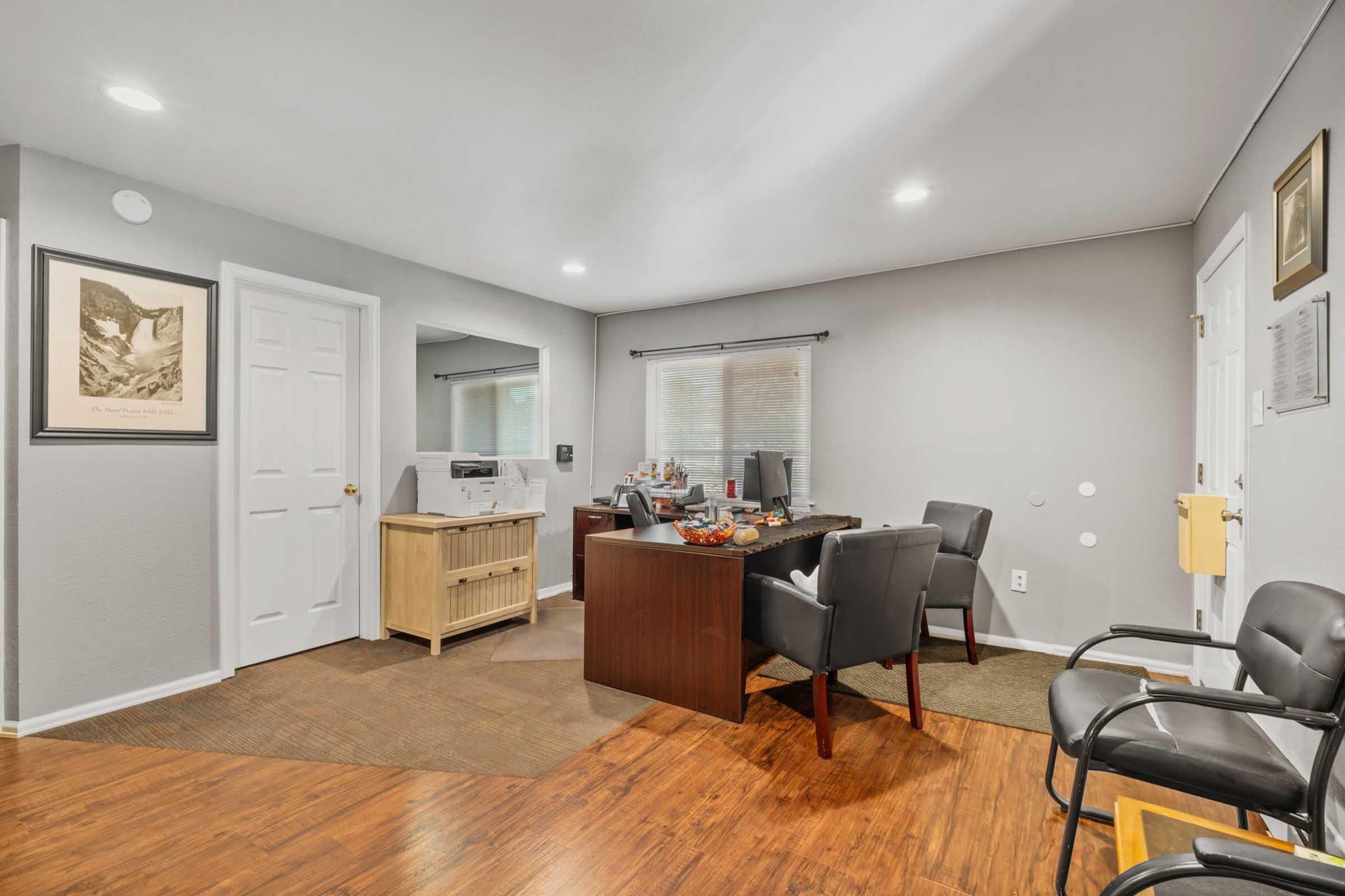 An office space featuring a desk with various items, two black chairs, a wooden cabinet, and a wall-mounted framed picture. The room has neutral wall colors and two light fixtures, providing a well-lit environment, with a window visible on one wall.