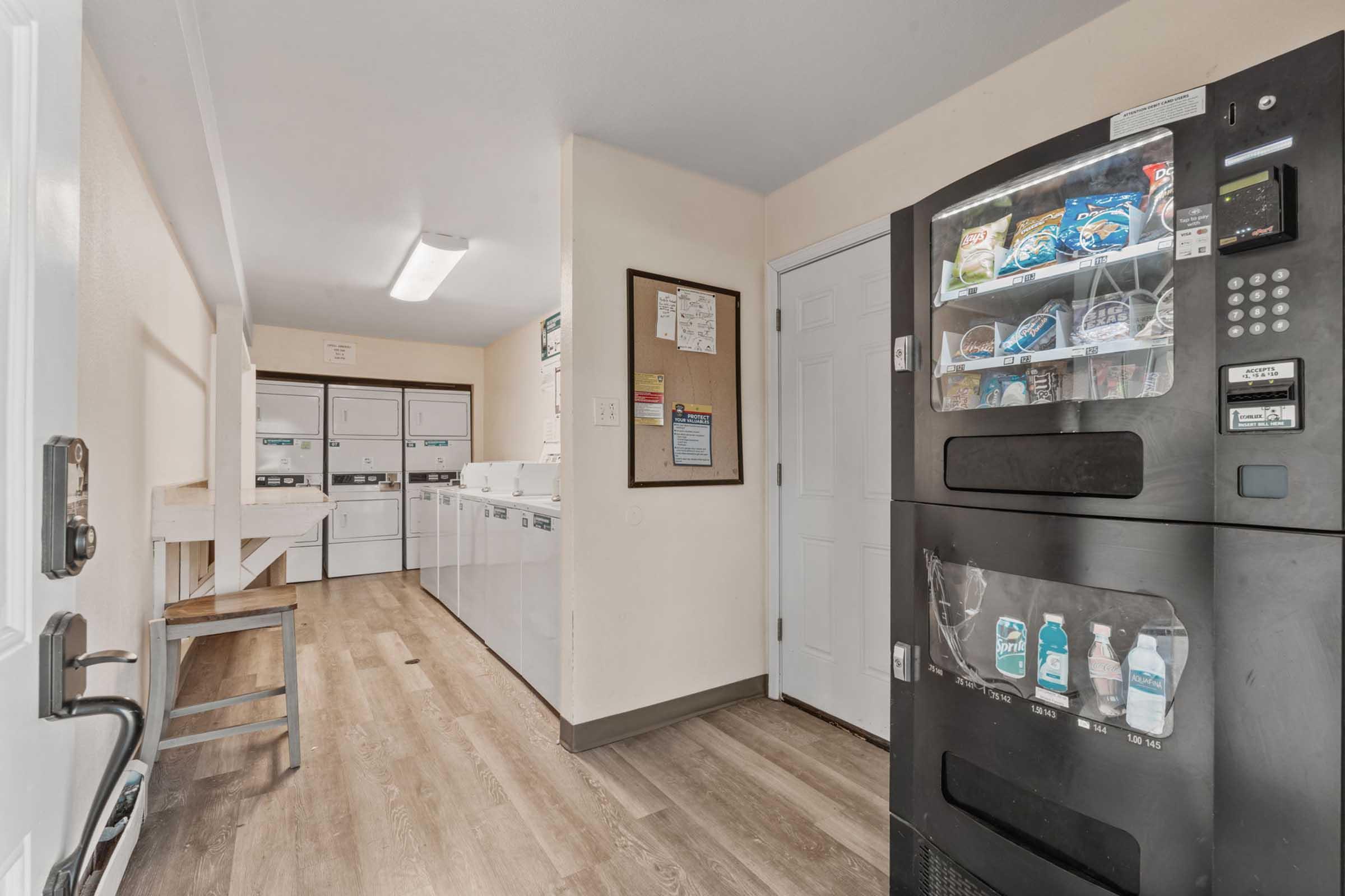 A laundry room featuring multiple washing machines and dryers against one wall. There is a bulletin board with notices and schedules. A vending machine, stocked with snacks and drinks, is positioned next to the door. The floor is wooden, and the lighting is bright, creating a clean and functional space.