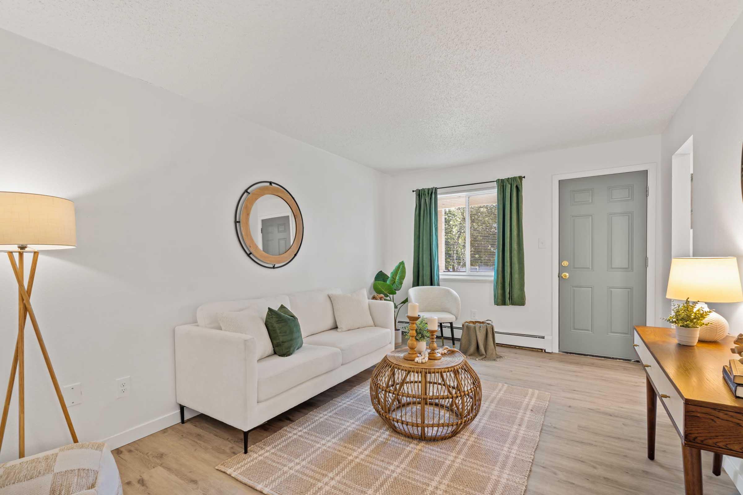A cozy living room featuring a white sofa with a green accent pillow, a round mirror on the wall, and a wooden coffee table. Natural light streams through a window adorned with green curtains, illuminating the space. A patterned area rug and potted plant add warmth, complemented by a floor lamp and a console table with decor items.