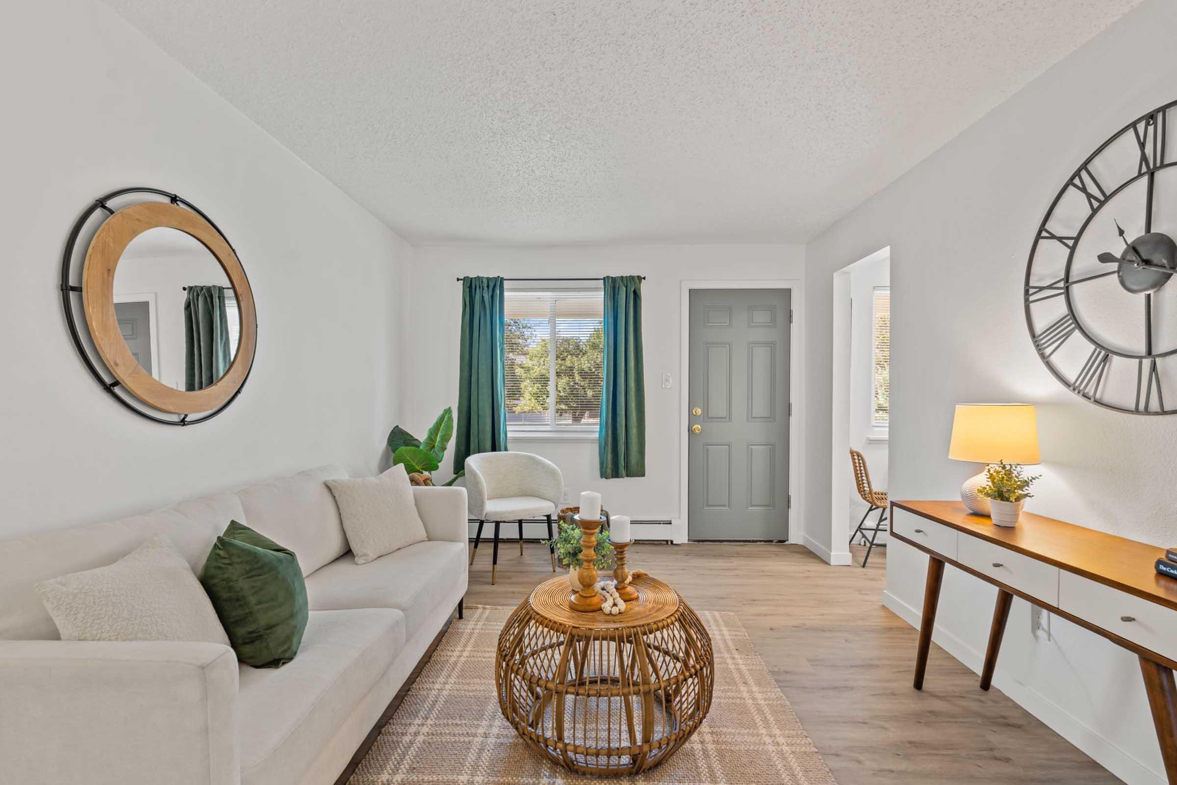 A cozy living room with light-colored walls and a textured ceiling. There is a small table with a woven design, a green plant, and a comfortable chair. To the right, a doorway leads to another room, while large windows provide natural light. A warm lamp adds a touch of ambiance.