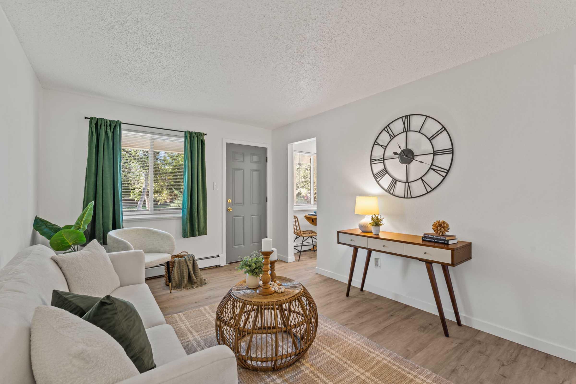 A cozy living room featuring a light-colored sofa, a round coffee table made of wicker, and a wooden console table with a lamp. The walls are painted white, and large windows with green curtains allow natural light in. A modern wall clock is displayed, enhancing the inviting atmosphere.
