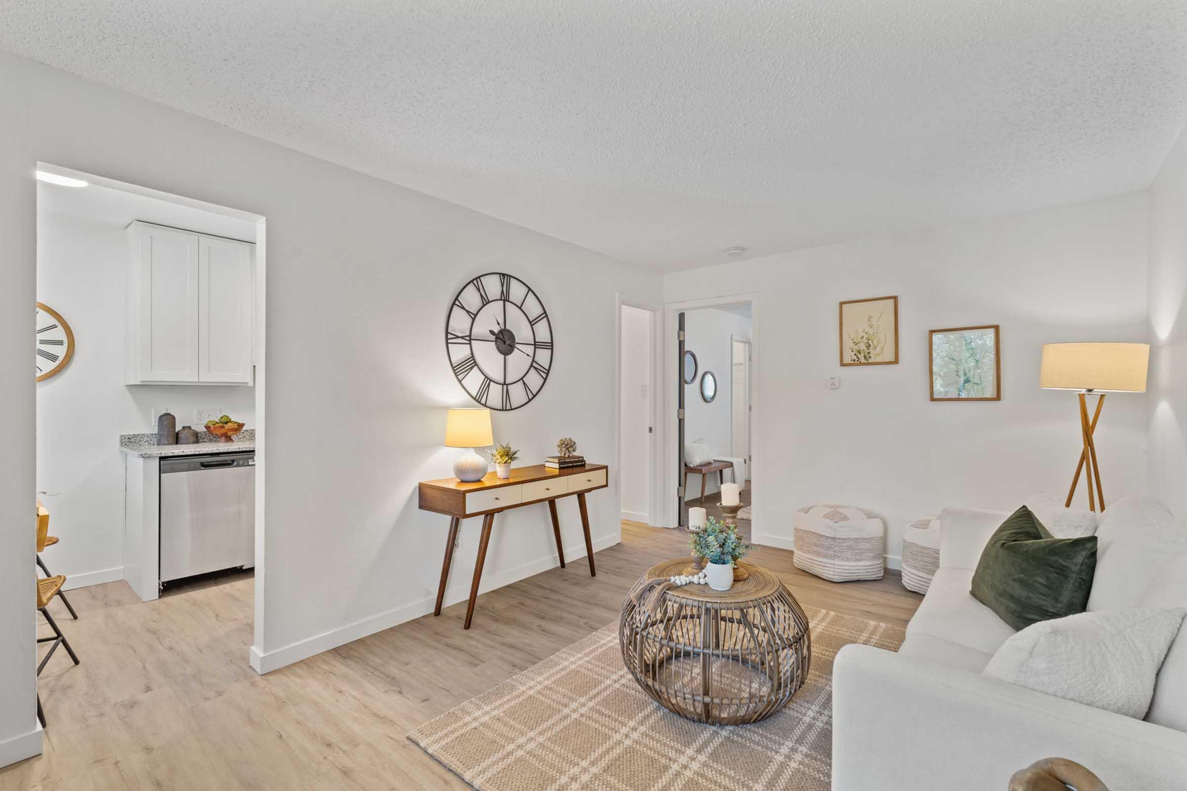 A cozy living room featuring a white sofa, a round coffee table made of woven material, and a large wall clock. There's a floor lamp beside the sofa, a textured area rug, and decorative items on the table. A glimpse of a kitchen is visible through an open doorway. The walls are painted in a soft, neutral color.