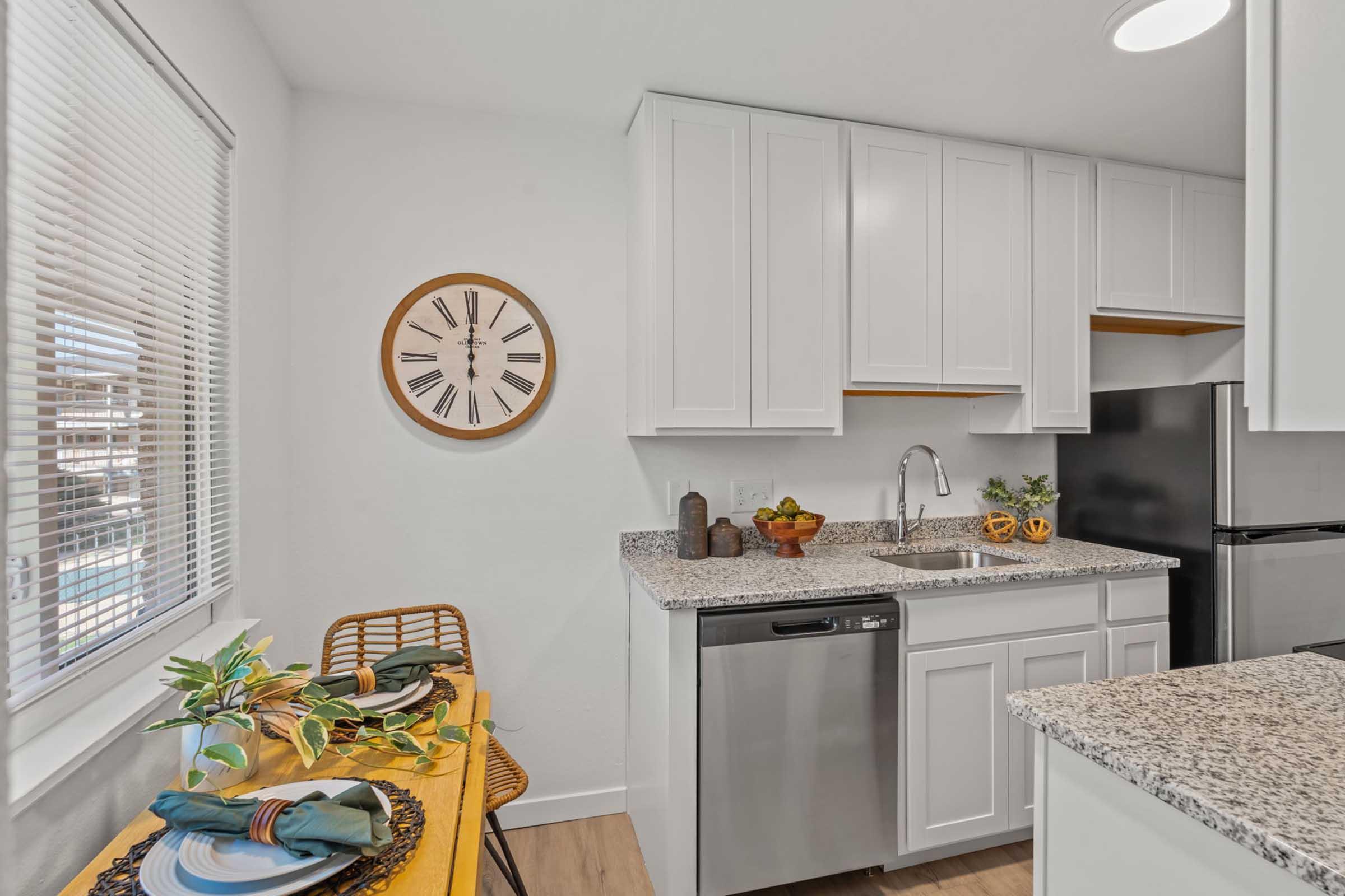 A modern kitchen featuring white cabinetry, a granite countertop, and stainless steel appliances. A round wall clock is mounted above the sink, and a small dining table with green napkins and decorative elements is set nearby, adding a touch of warmth to the space. Natural light filters through the window.