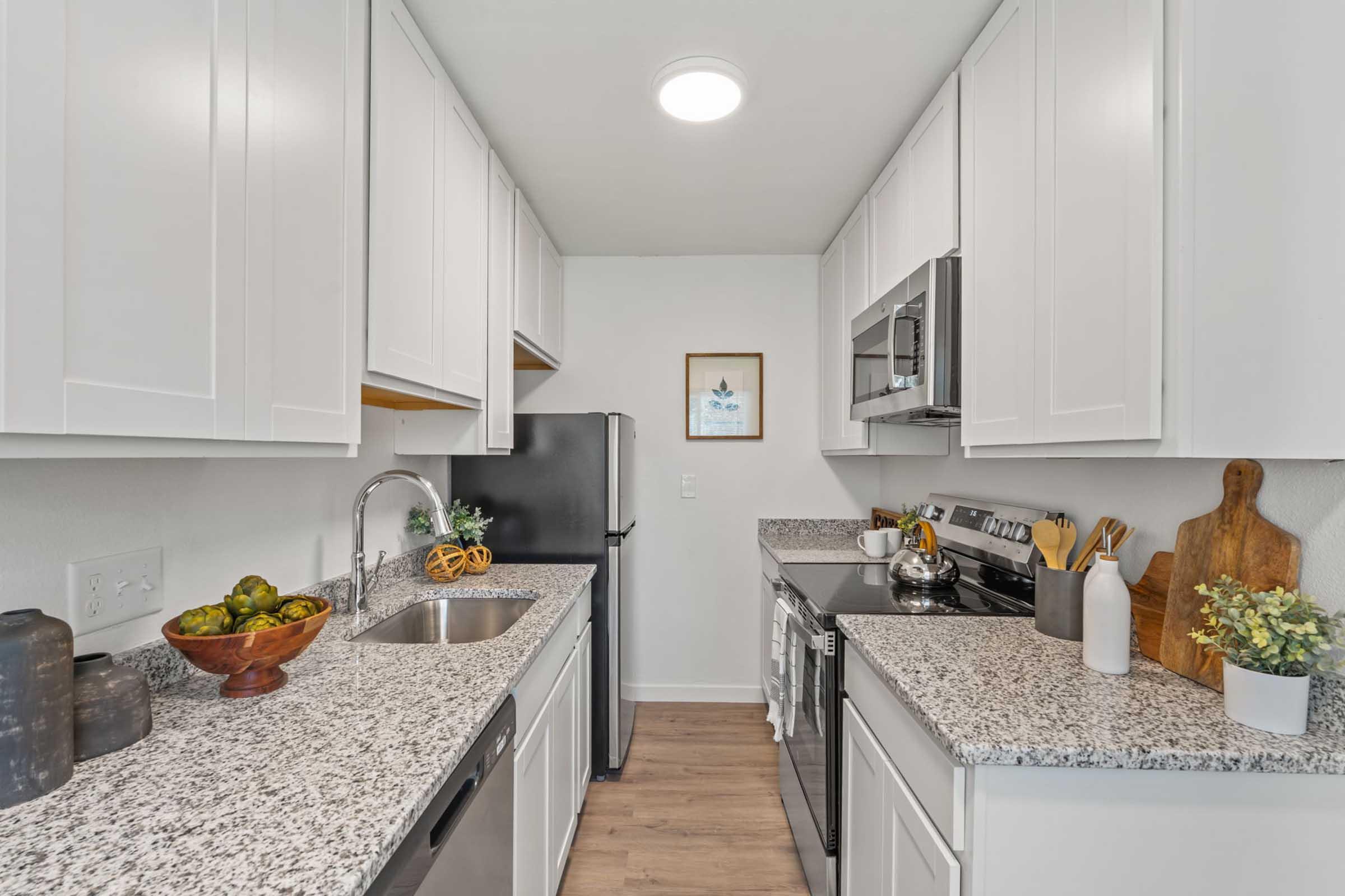 Modern kitchen featuring white cabinets, a black refrigerator, stainless steel oven and microwave. The countertop is made of granite, adorned with a wooden bowl of fruit, a potted plant, and kitchen utensils. Bright overhead lighting enhances the spacious feel of the area.
