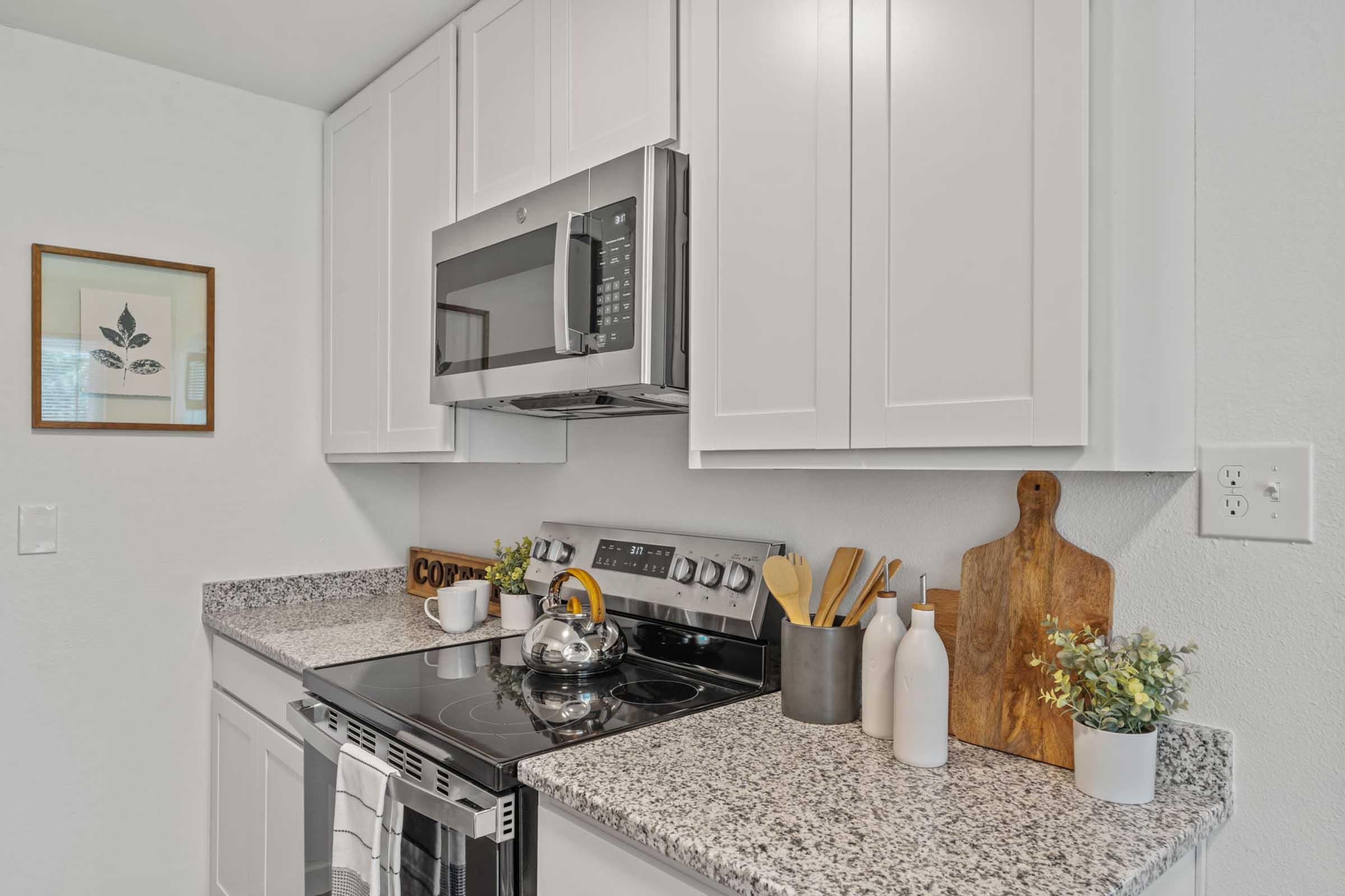A modern kitchen with white cabinets, a stainless steel microwave above a stove, and granite countertops. On the counter, there is a kettle, wooden cutting board, assorted utensils in a holder, and decorative bottles with a small plant. A framed botanical print is visible on the wall.