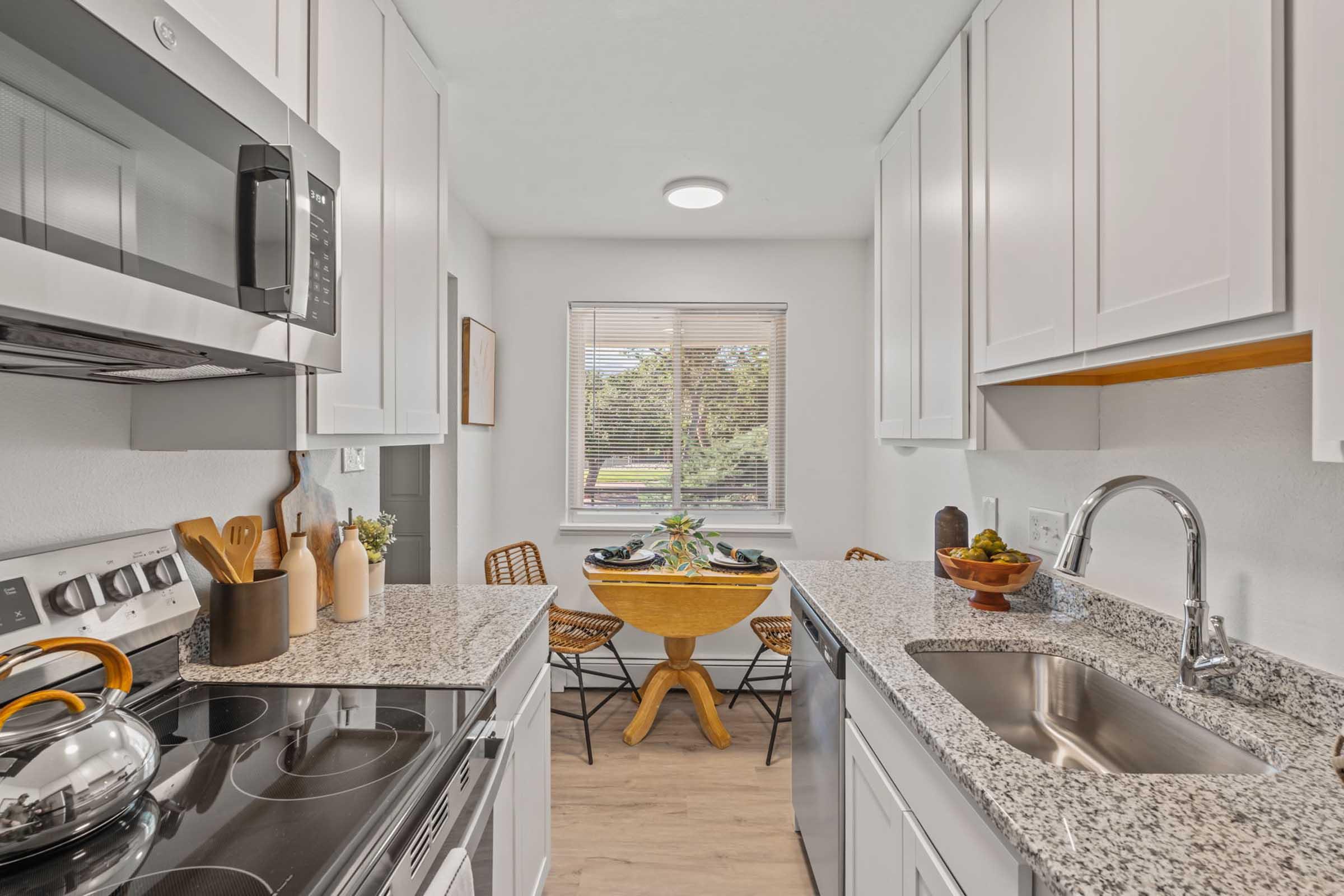 A modern kitchen featuring white cabinets, granite countertops, and a stainless steel sink. An electric stove and microwave are visible, along with wooden utensils and a kettle. A round wooden dining table with chairs is situated near a window, allowing natural light to fill the space.