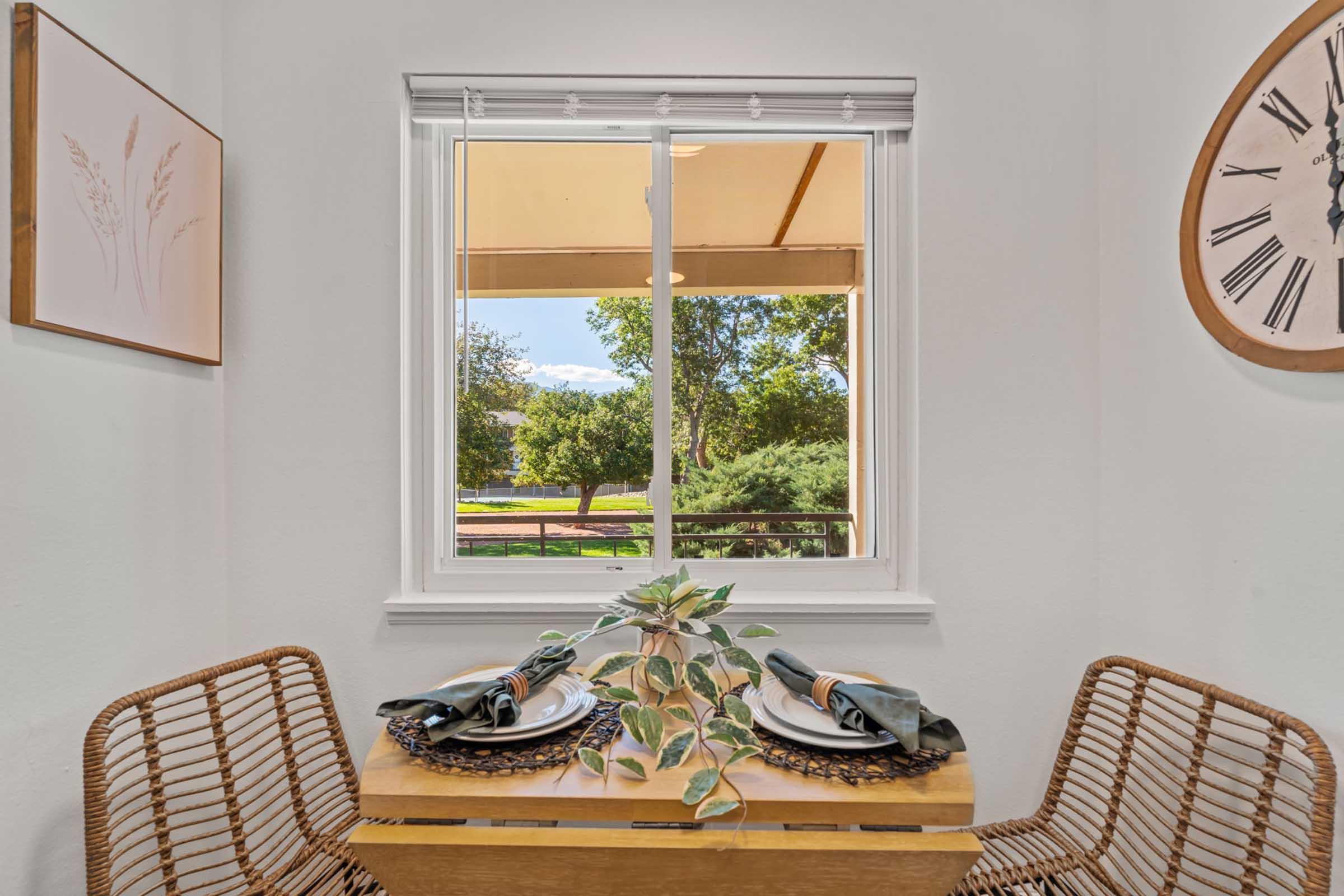 A small dining area with a wooden table set for two, featuring green napkins and decorative plates. The window offers a view of trees and a sunny outdoor space, framed by light-colored walls and decorative wall art, including a clock and framed botanical illustration.