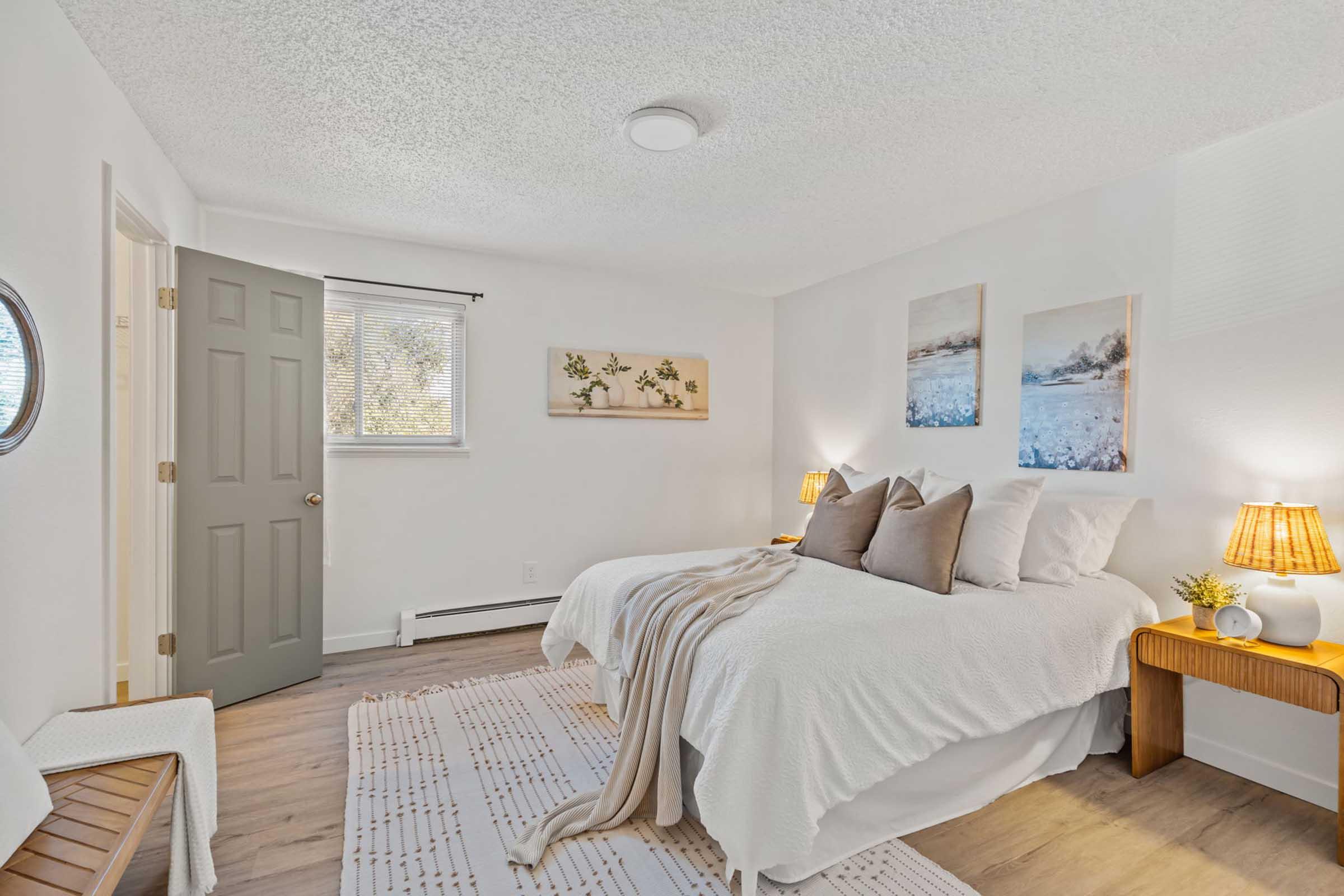 A cozy bedroom featuring two beds with white bedding, decorative pillows, and bedside tables with lamps. The room has light-colored walls, a window with sheer curtains, and artwork depicting serene landscapes. A door leads to the outside, and a textured rug adds warmth to the space.