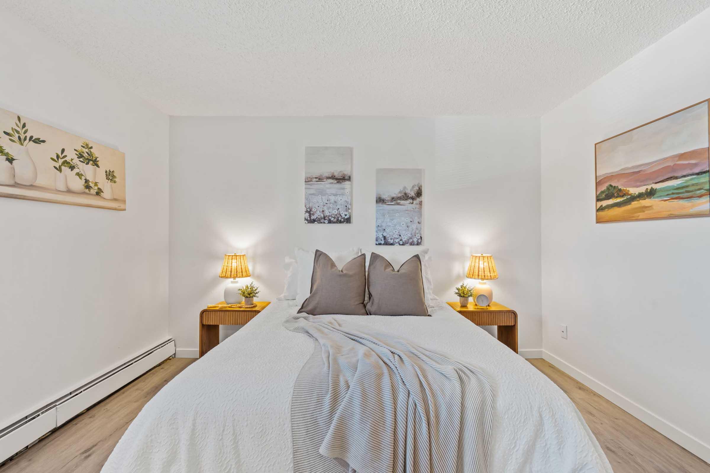 A cozy bedroom featuring a neatly made bed with gray pillows and a light blanket. On either side of the bed, there are wooden nightstands with lamps. The walls are adorned with three artworks depicting serene landscapes, contributing to a calm and inviting atmosphere. Natural light illuminates the space.