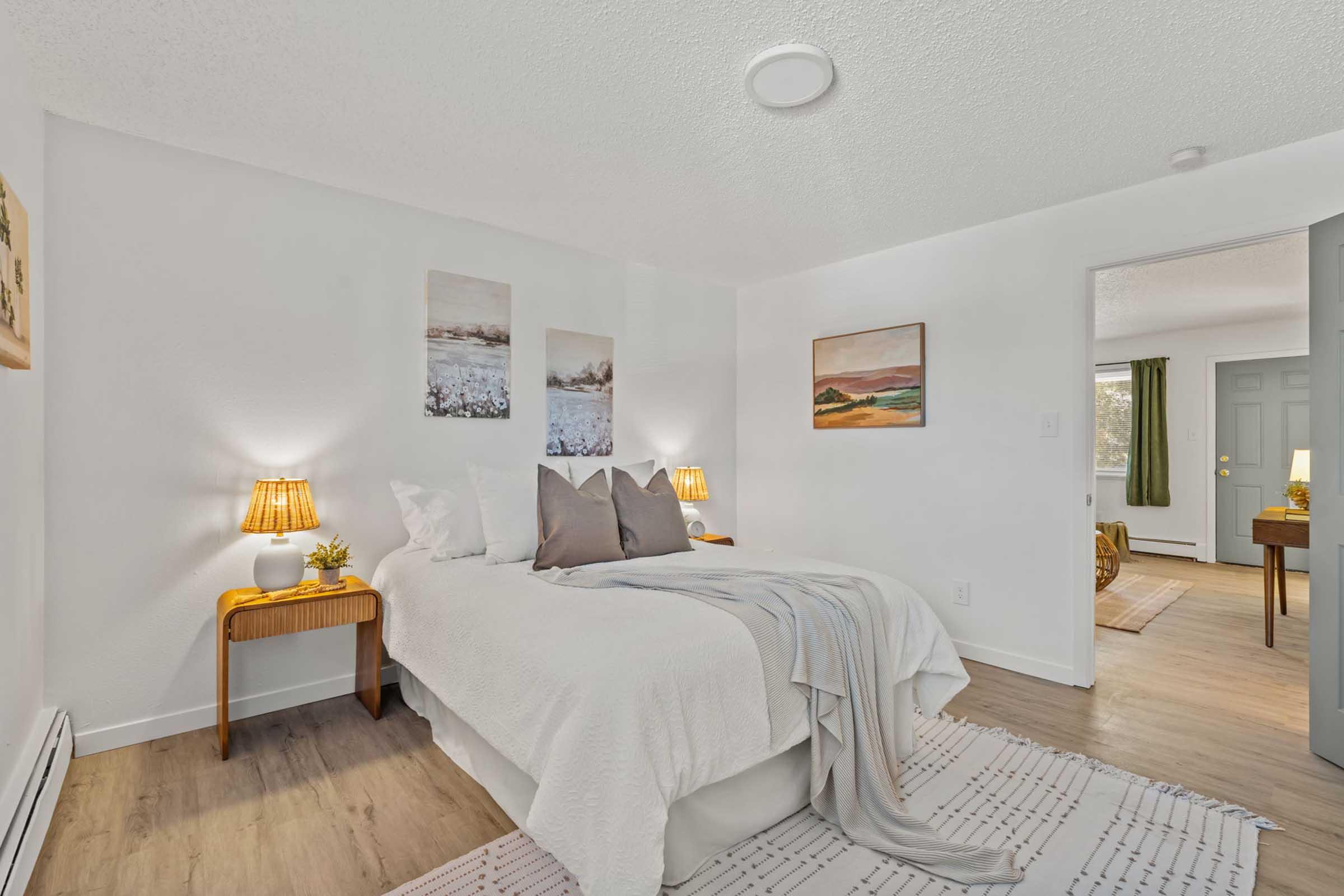 A cozy bedroom featuring a bed with light-colored bedding and decorative pillows, two bedside tables with lamps, and wall art depicting serene landscapes. The room has neutral walls and flooring, with a rug and a glimpse of another room through an open door. Natural light filters in, creating a warm atmosphere.