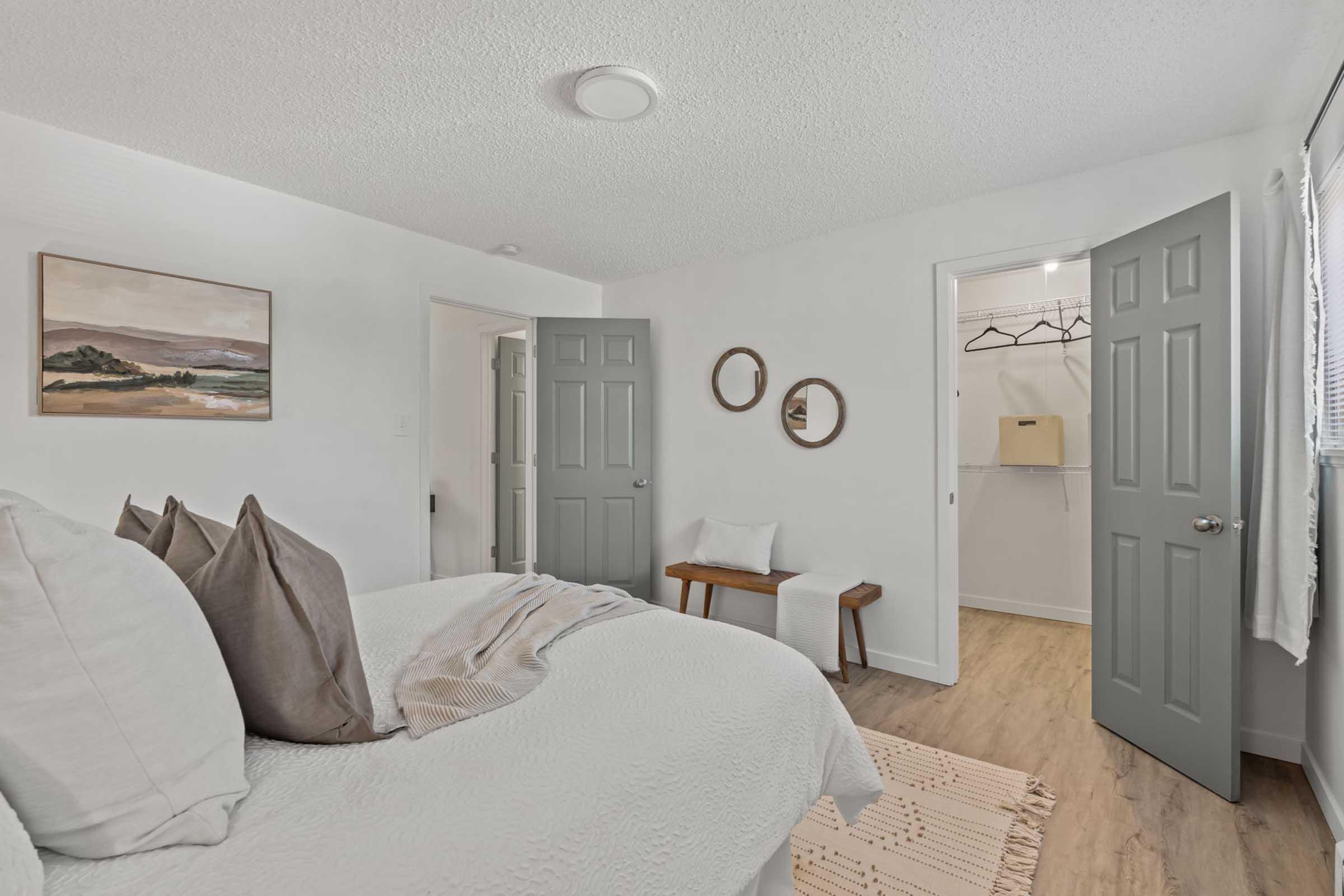 A cozy bedroom featuring a bed with cream bedding and decorative pillows. There are two round mirrors on the wall, a wooden bench, and an open closet with hangers. A painting of a landscape is displayed above the bed, and the room has light-colored walls and gray doors. Natural light illuminates the space.