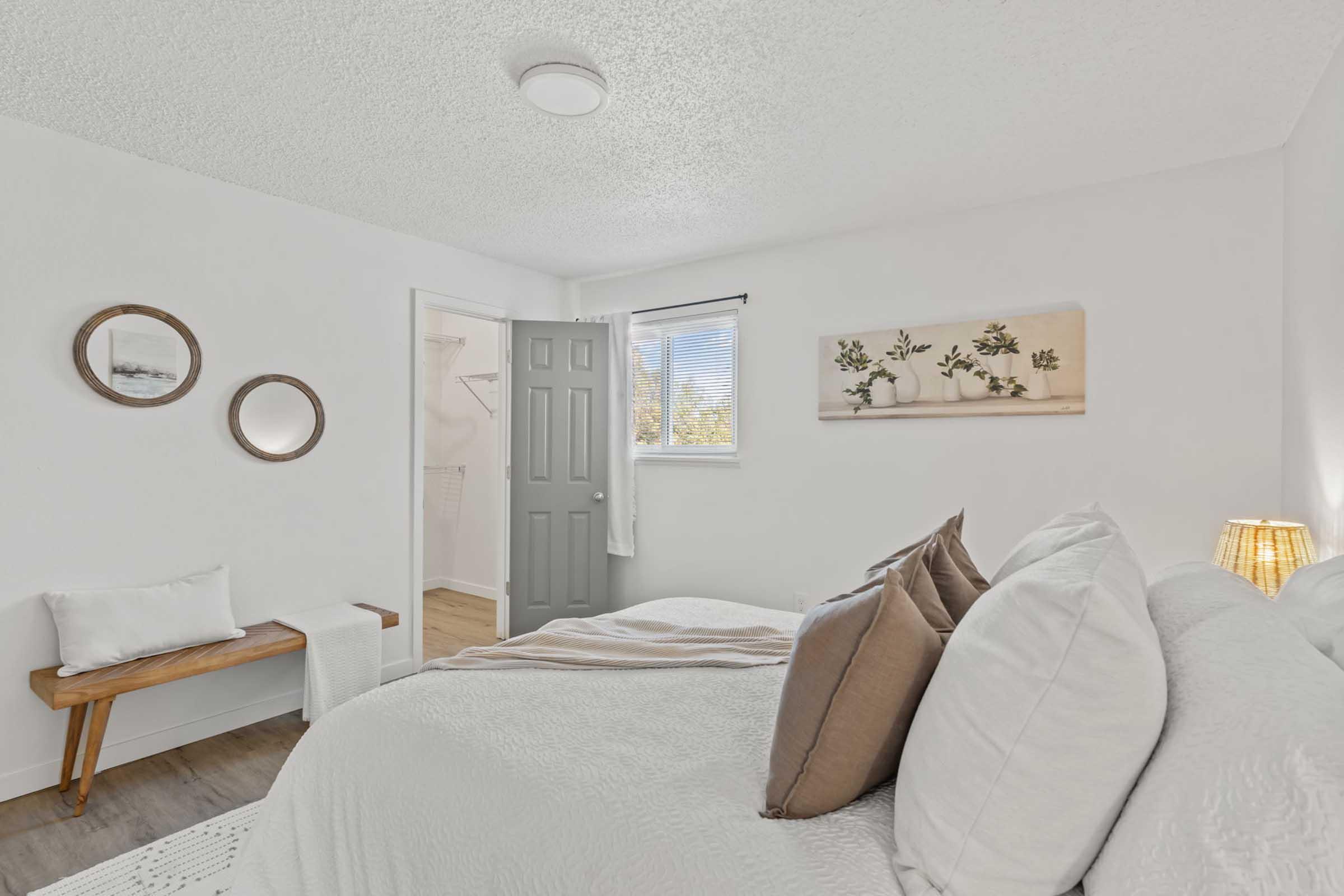 A cozy bedroom featuring a neatly made bed with decorative pillows, a small wooden bench with a cushion, and two round mirrors on the wall. A door leads to a small closet space, and a piece of wall art depicting plants adds a touch of decor. Natural light filters in through a window, enhancing the inviting atmosphere.