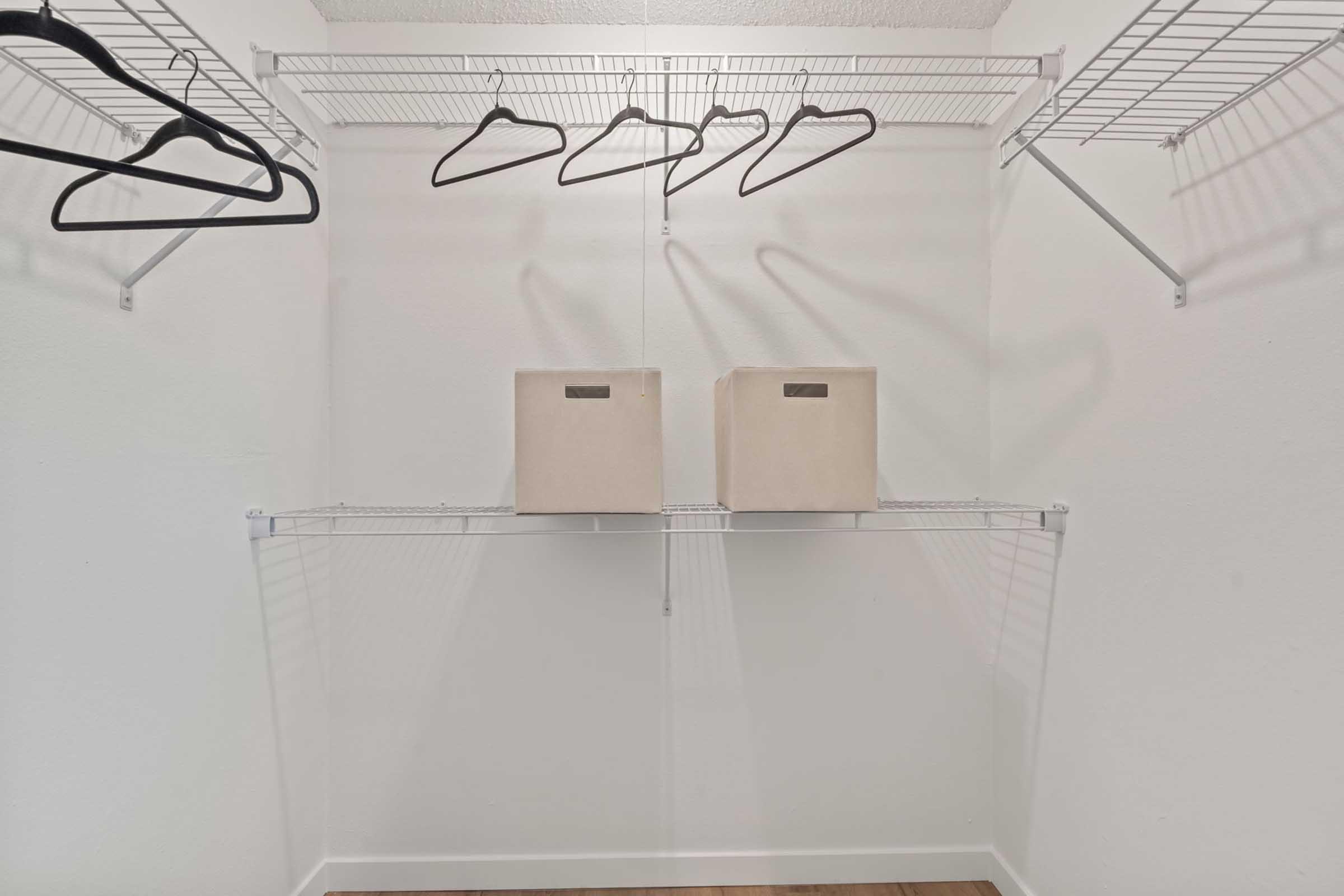 A minimalist closet with wire shelves. Two beige storage boxes are placed on the lower shelf, and several black hangers are hanging from the upper shelves. The walls are white, and the floor is wooden, creating a clean and organized space.