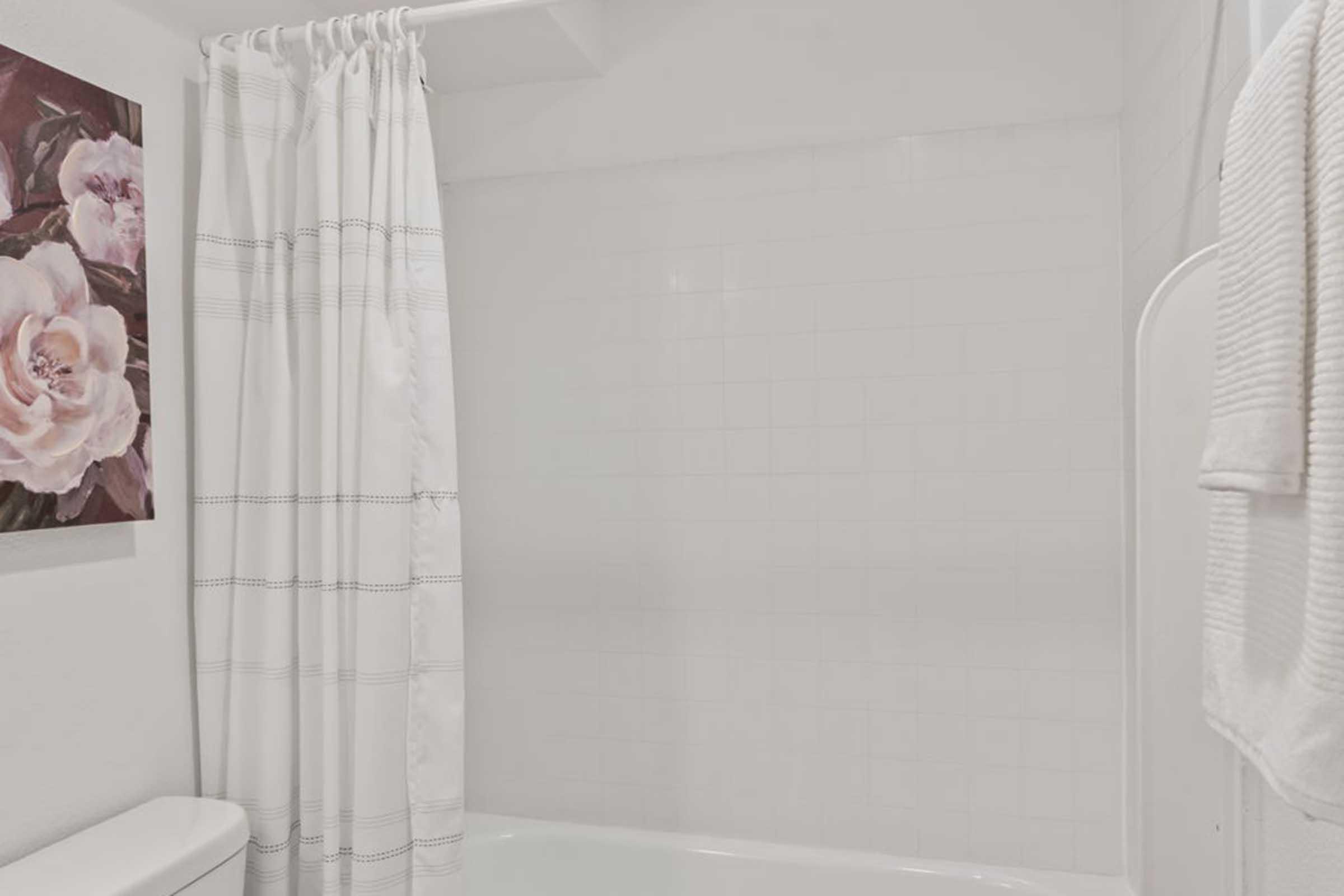 A clean and minimalist bathroom featuring a shower with a white curtain. To the left, a floral artwork is displayed on the wall, and a neatly folded white towel hangs nearby. The overall color scheme is light and bright, contributing to a fresh and airy atmosphere.