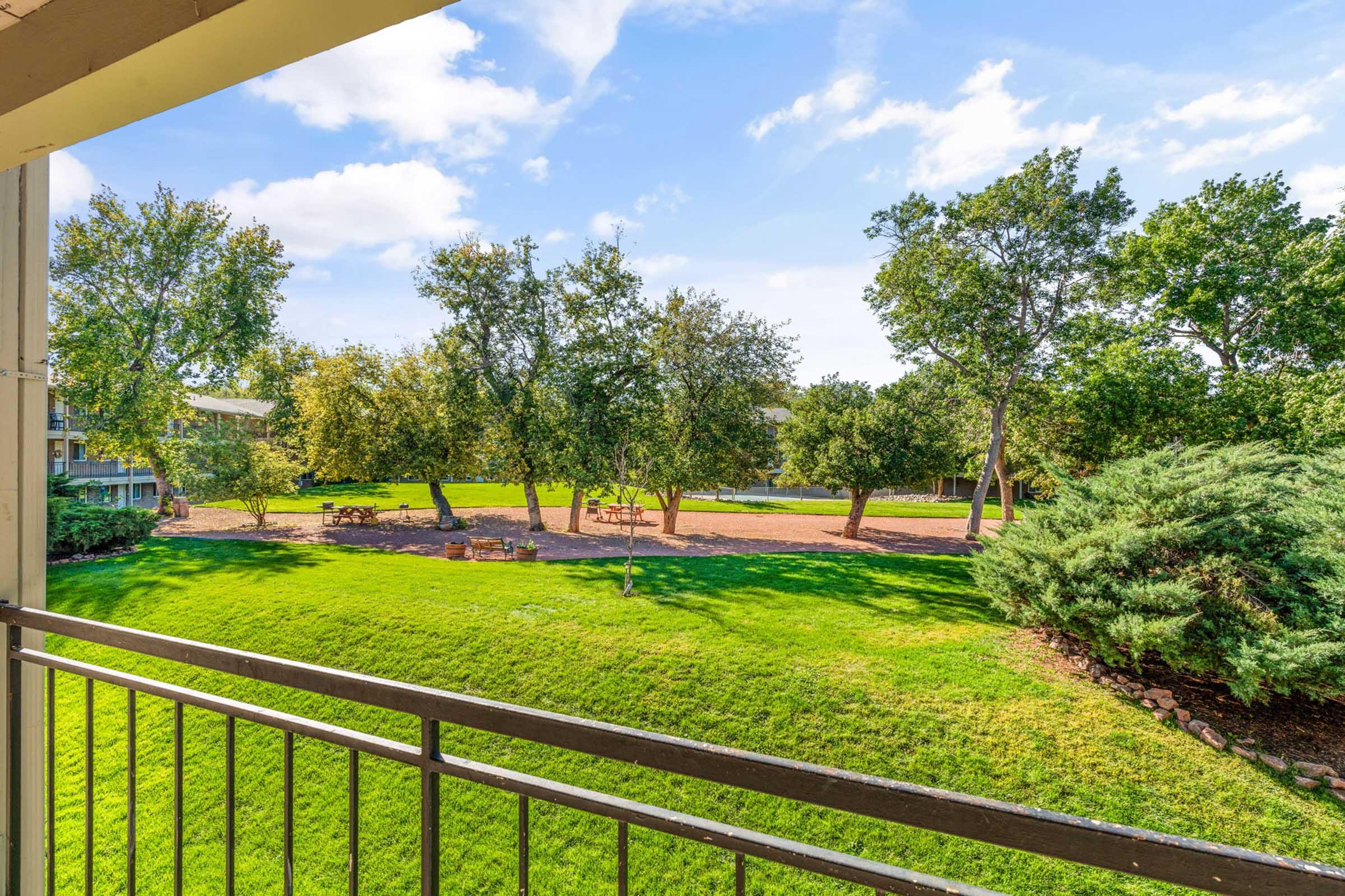 View from a balcony overlooking a lush green area with trees and picnic tables. The landscape features well-maintained grass and a serene atmosphere, with blue sky and fluffy clouds in the background.