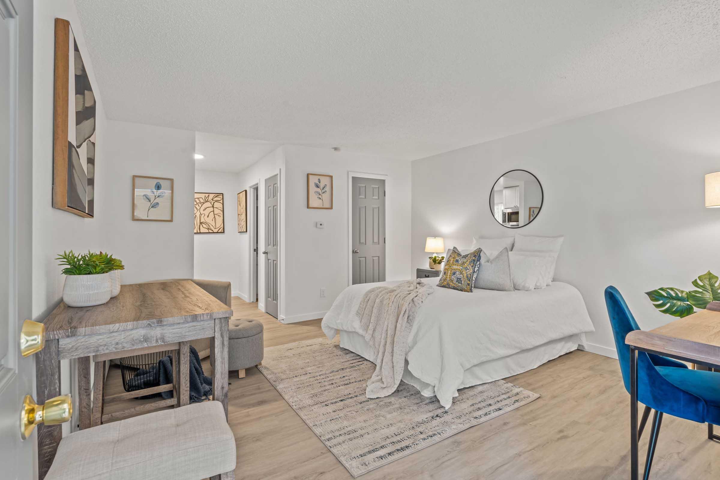 A cozy, modern studio apartment featuring a large bed with decorative pillows, a round mirror on the wall, a small desk and chair, and a dining area with a table and blue chairs. Light-colored walls and wooden flooring create a bright and inviting atmosphere. Decorative plants and wall art enhance the decor.