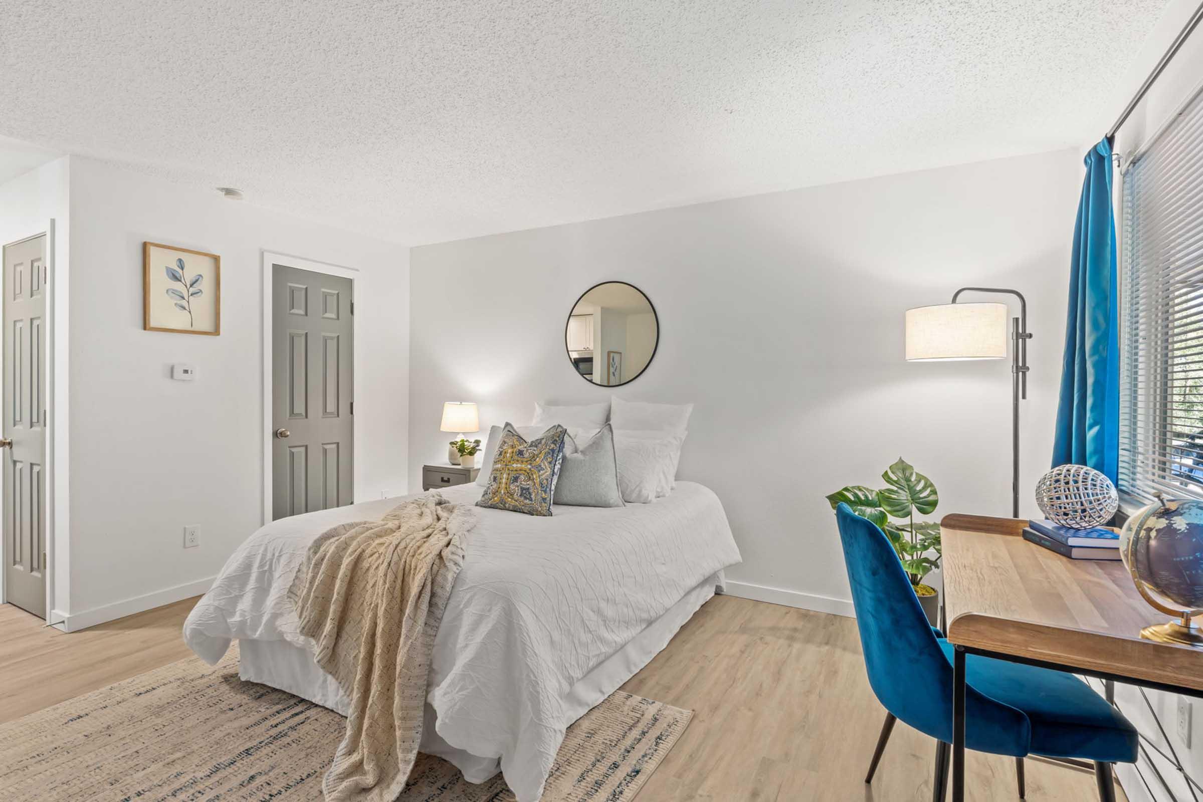 Bright and inviting bedroom featuring a bed with decorative pillows and a cozy throw. A round mirror hangs above a nightstand with a lamp. A desk and chair are positioned near a window, complemented by a green plant and a globe. Light-colored walls and wooden flooring enhance the airy feel of the space.